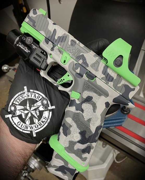 A person holding a handgun with a custom camouflage pattern in shades of gray, black, and white, accented with bright green parts including the trigger, slide, and grips. The person is wearing a black glove with a logo from Apex Tactical and is in a workshop or gun shop environment.