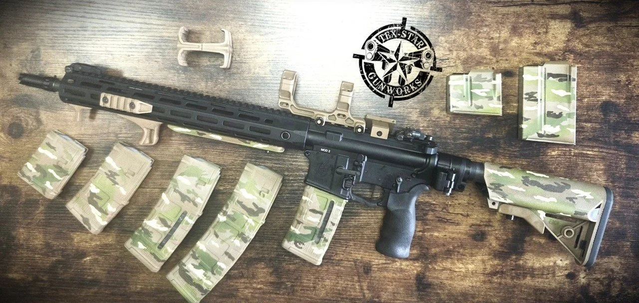 An assault rifle with camouflage-patterned magazines and accessories laid out on a wooden surface.