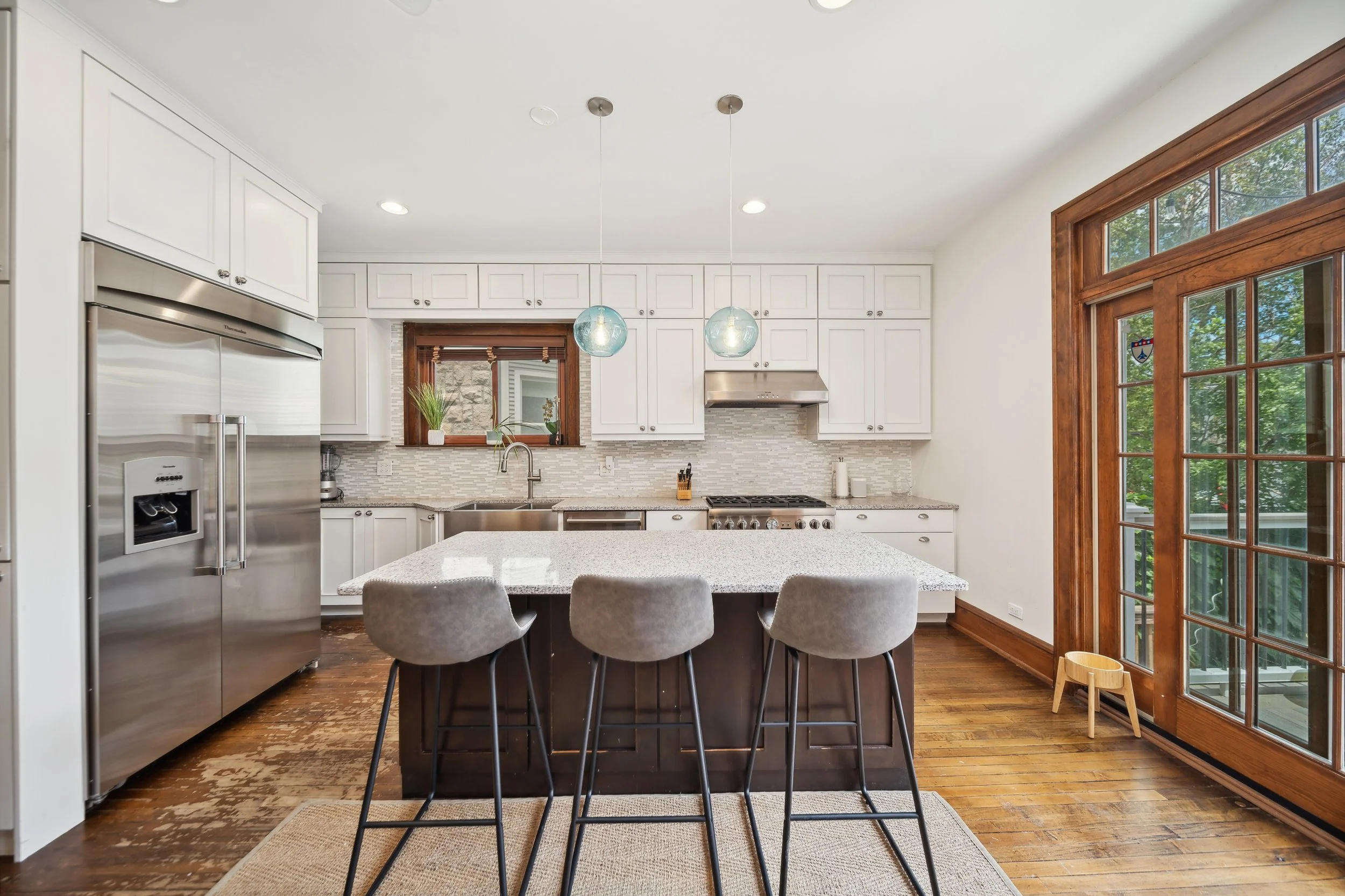 Modern kitchen with white cabinets, granite countertops, and stainless steel appliances, including a refrigerator and stove, with a large wooden-framed glass door on the right and a small window above the sink, featuring a kitchen island with four high chairs.