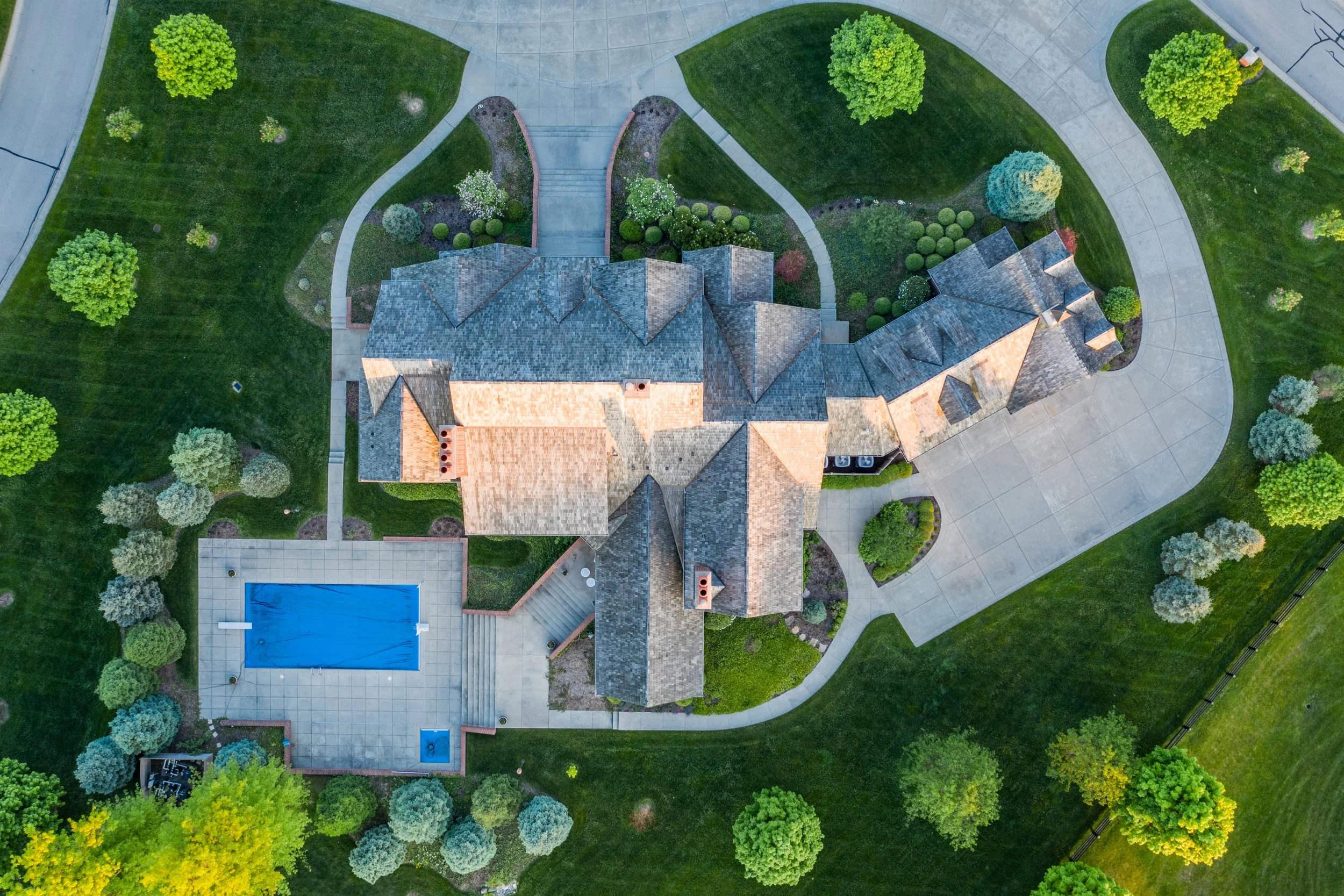 Aerial view of a large house with a grey shingle roof, a swimming pool, and landscaped yard with trees and pathways.