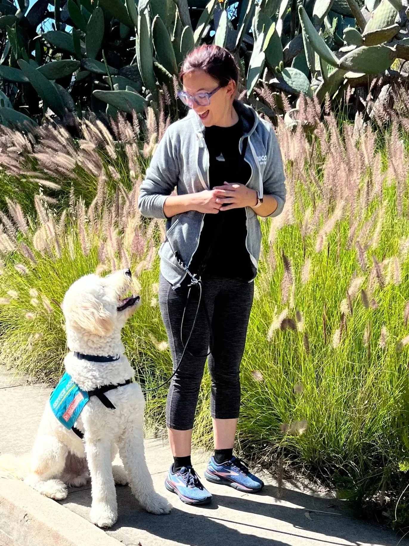 A golden doodle service dog in training looks up and smiles at his trainer who is a woman wearing a jogging outfit and smiling back at him