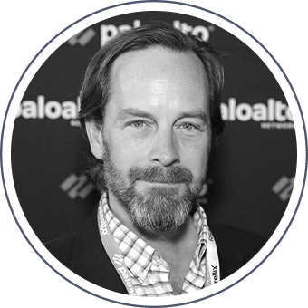 Black-and-white headshot of a man with a beard, wearing a blazer over a checkered shirt and a lanyard