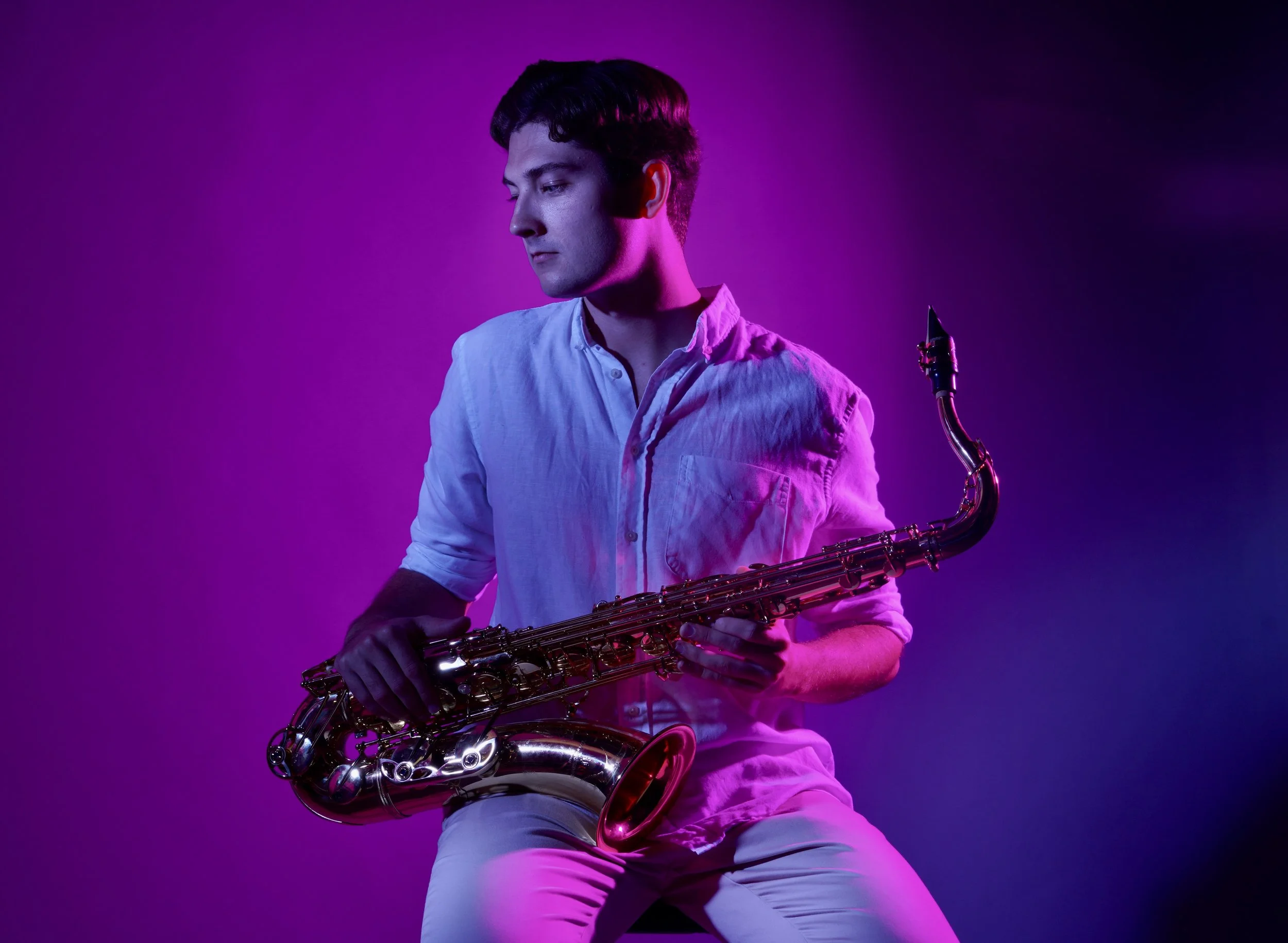 A young man with dark hair wearing a light-colored button-up shirt, holding a saxophone against a purple background with pink lighting.