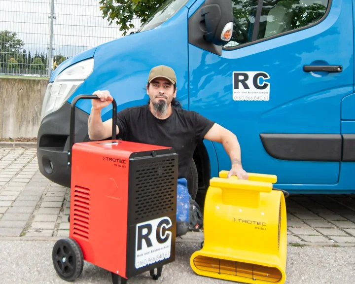 Ein Mann sitzt auf dem Boden neben einem blauen Fahrzeug mit einem RC-Logo auf der Tür. Er hat eine grau-braune Kappe auf und hält eines der Geräte in der Hand. Vor ihm stehen eine rote und eine gelbe Reinigungsmaschine.