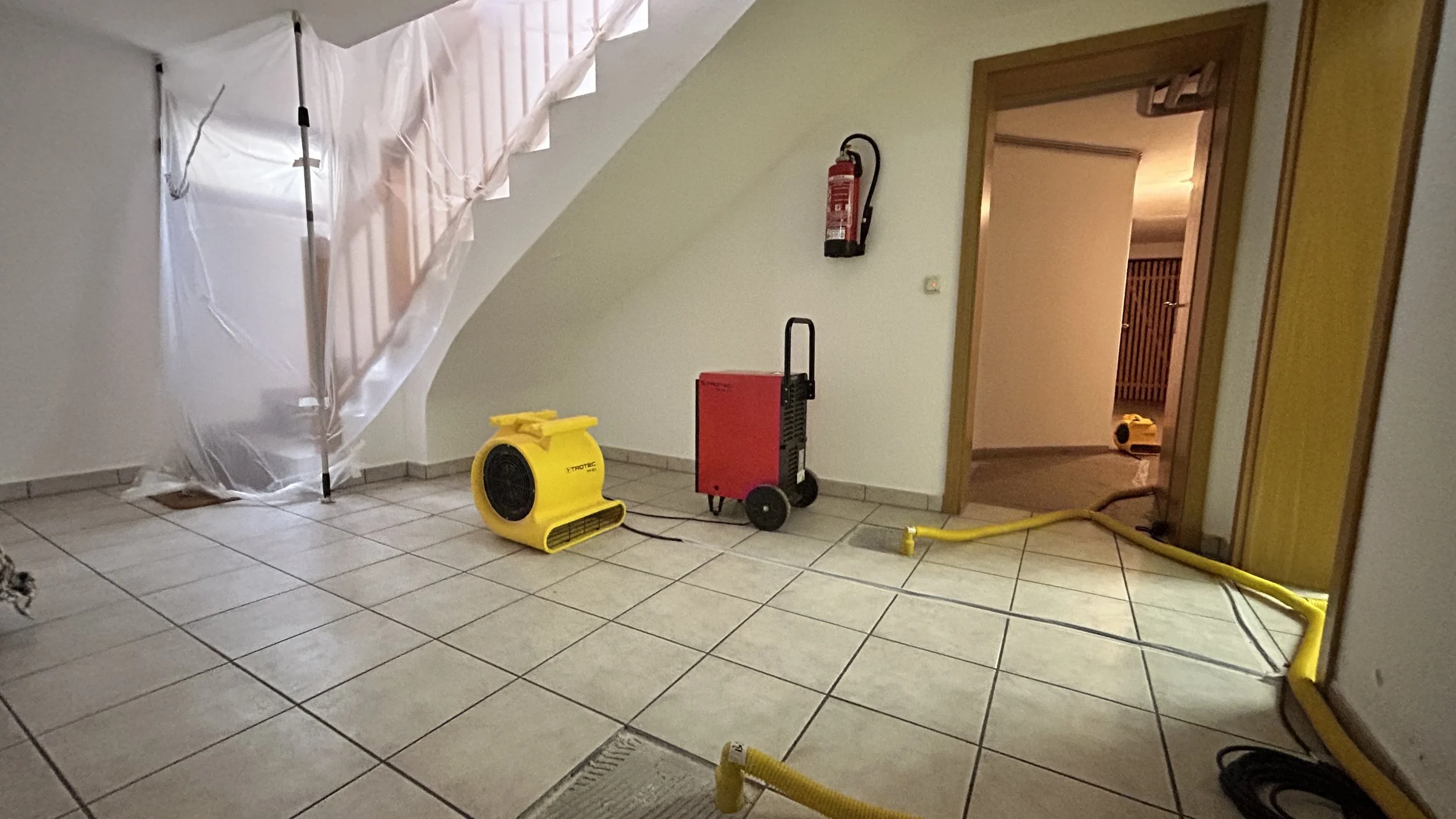 Innenaufnahme eines Raumes mit Reinigungsgeräten, darunter ein Ventilator, ein Heizgerät, ein Geräteschrank, eine Tür, ein Feuerlöscher an der Wand, eine gelbe Überwachungskamera und eine gelbe Schläuche. Im Hintergrund sind Bauplanen und Handwerkerarbeiten sichtbar.