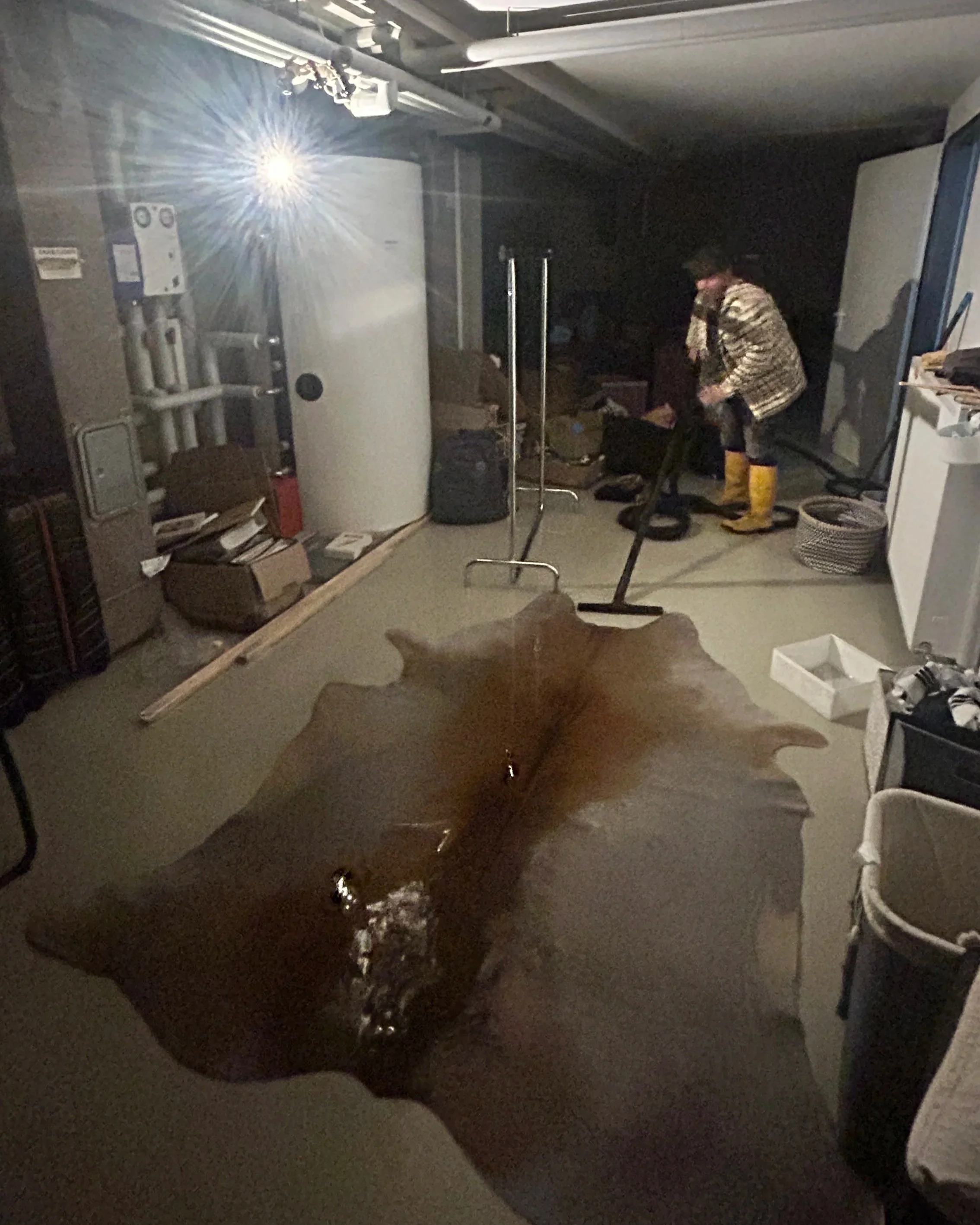 Eine Person im Raum trägt Gummistiefel und eine Jacke, während sie sich vor einem schwarzen Wasserbecken befindet. Es gibt verschiedene Gegenstände wie eine Wasserboiler, Kartons, einen Besen und andere Haushaltsgegenstände im Raum.