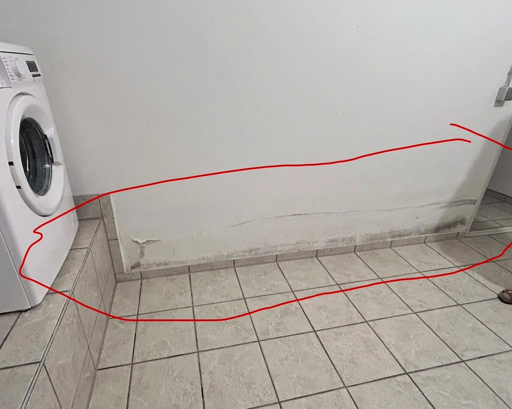 Waschmaschine neben einer verschmutzten Wand, deren unterer Bereich mit Schimmel oder Schmutz bedeckt ist, in einem Raum mit Fliesenboden.