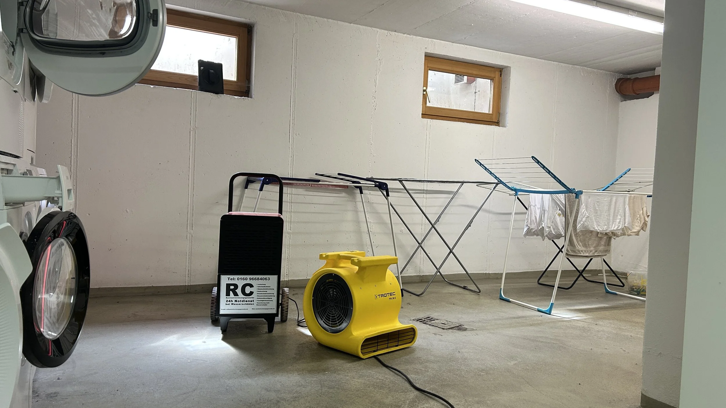 Waschraum mit Waschmaschinen, Trockner, Wäscheleinen, einem Ventilator, einem Wasserschneidetisch, Fenstern, einem Heizkörper und Reinigungsausrüstung.