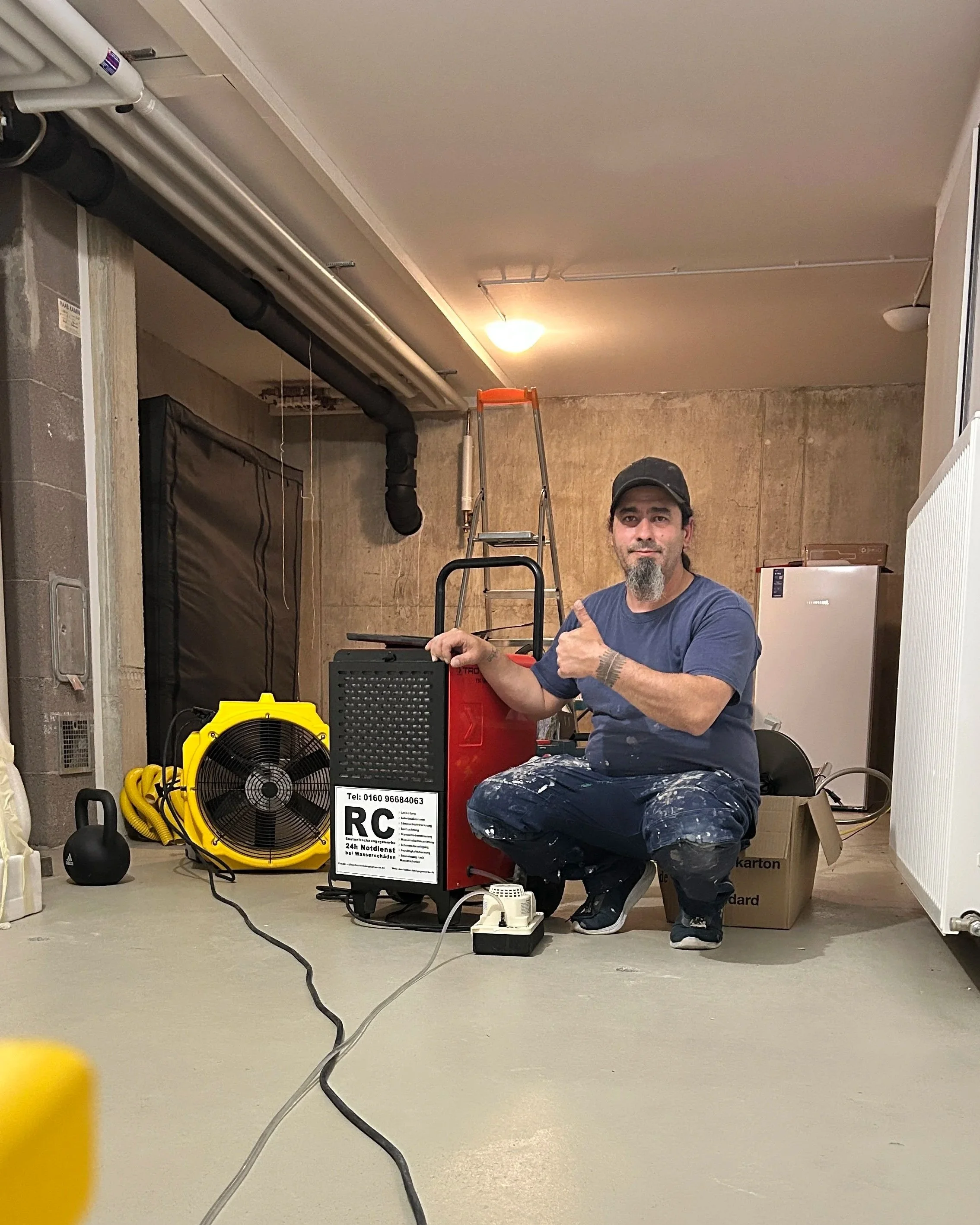 Ein Mann mit dunklem Hut und Bart sitzt in einem Raum voller technischer Geräte und Equipment. Im Vordergrund befinden sich Kabel, ein Ventilator sowie eine zentrale Wasserpumpe. Der Raum hat ungeputzte Wände und es ist eine Leiter im Hintergrund sichtbar.
