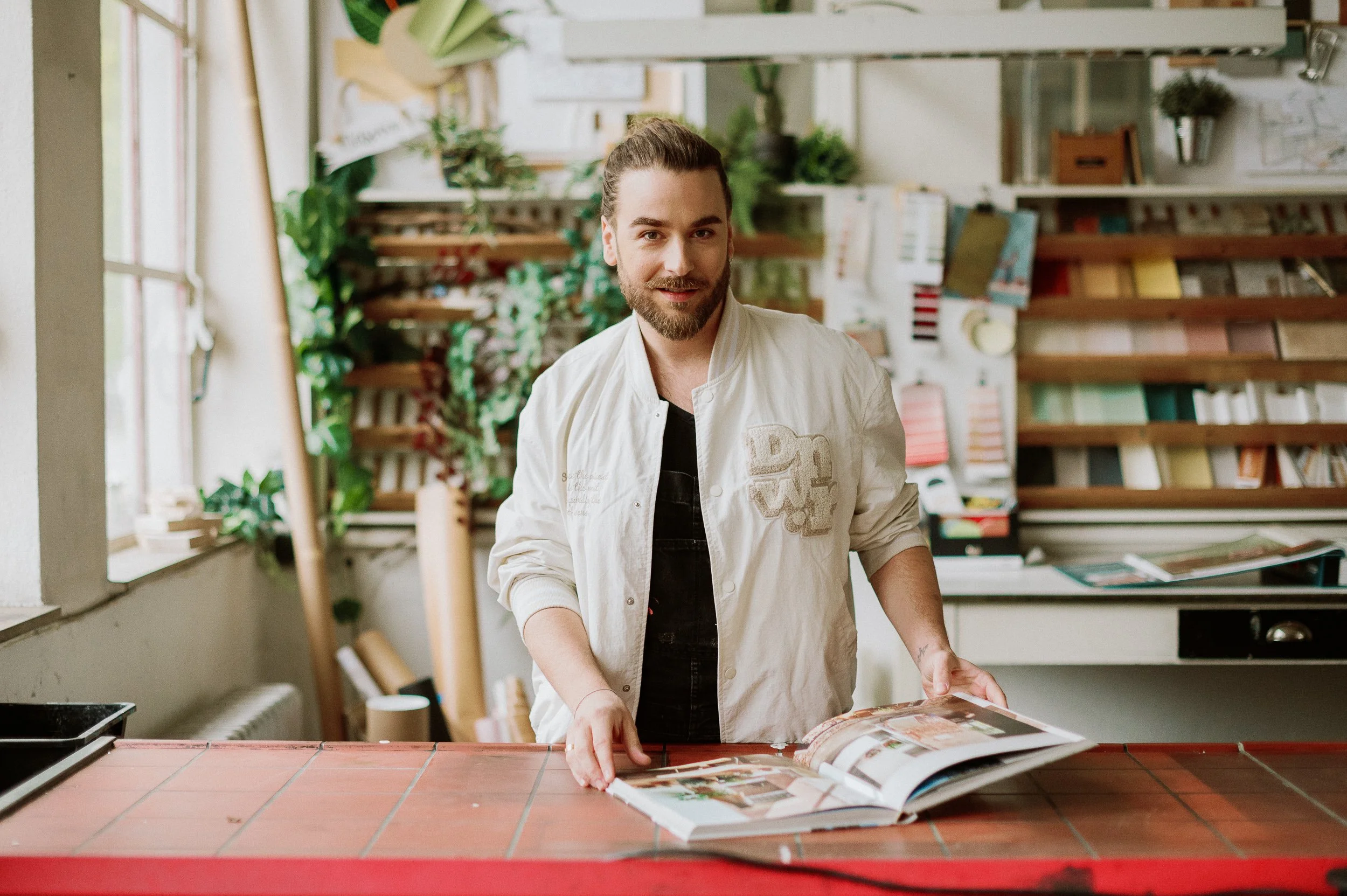 Ein Mann mit braunem Haar und Bart, trägt eine weiße Jacke, steht hinter einem roten Tresen in einem Laden, liest ein offenes Magazin, im Hintergrund Regale mit Bändern und Dekorationsartikeln, viel Grünpflanzen