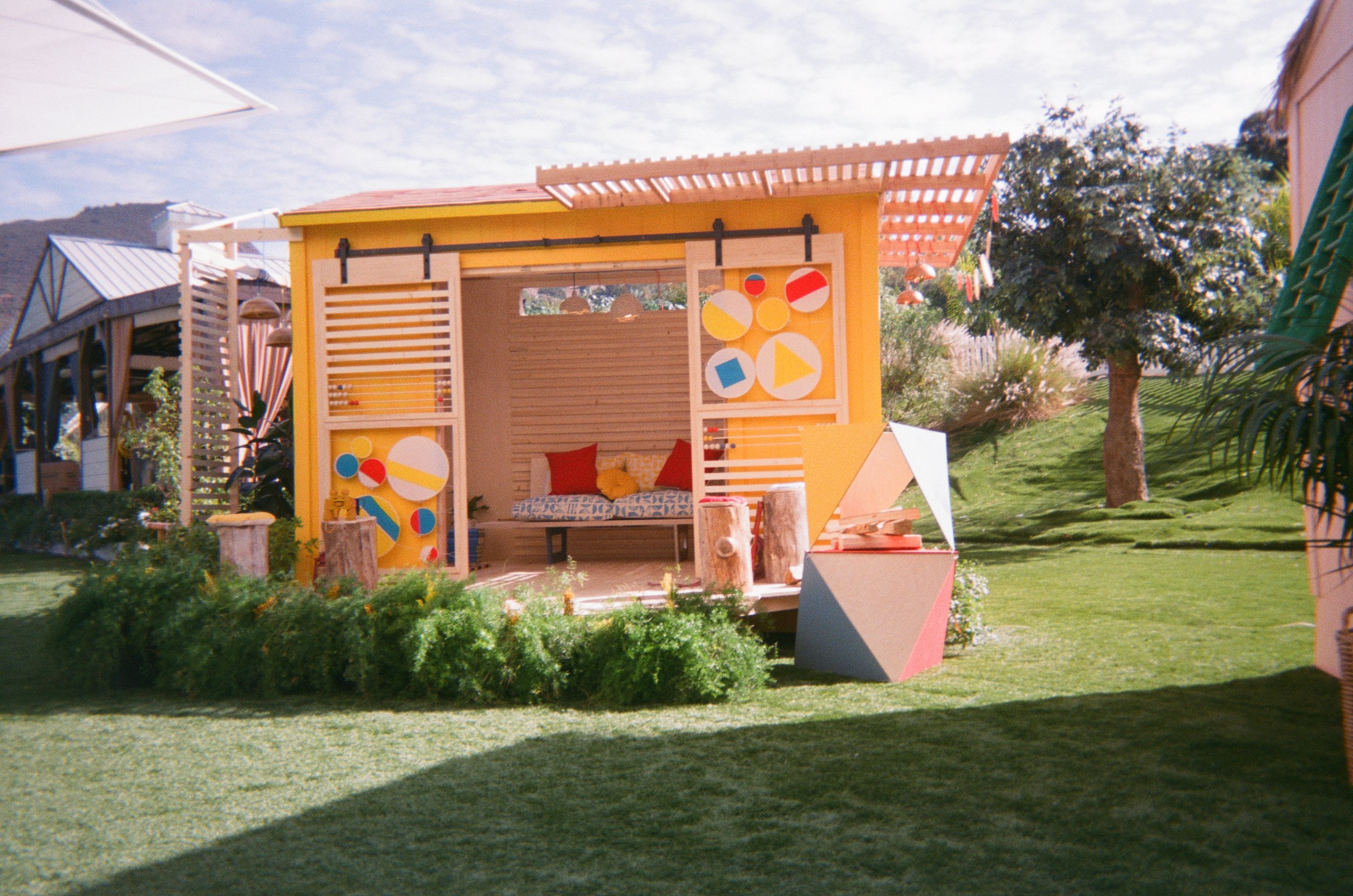A small yellow outdoor structure with a shaded roof, decorated with colorful geometric shapes, and featuring a small bed or seating area inside, situated on a well-maintained grassy lawn with trees and neighboring buildings in the background.