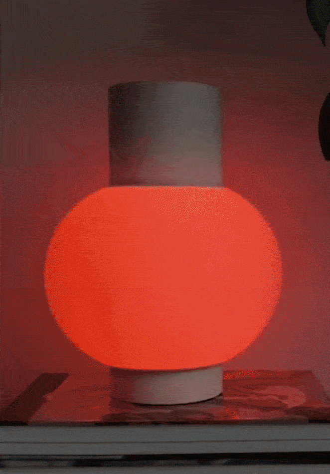 A lamp with a glowing orange-red rounded shade and a white base, sitting on a wooden surface with a coaster underneath.