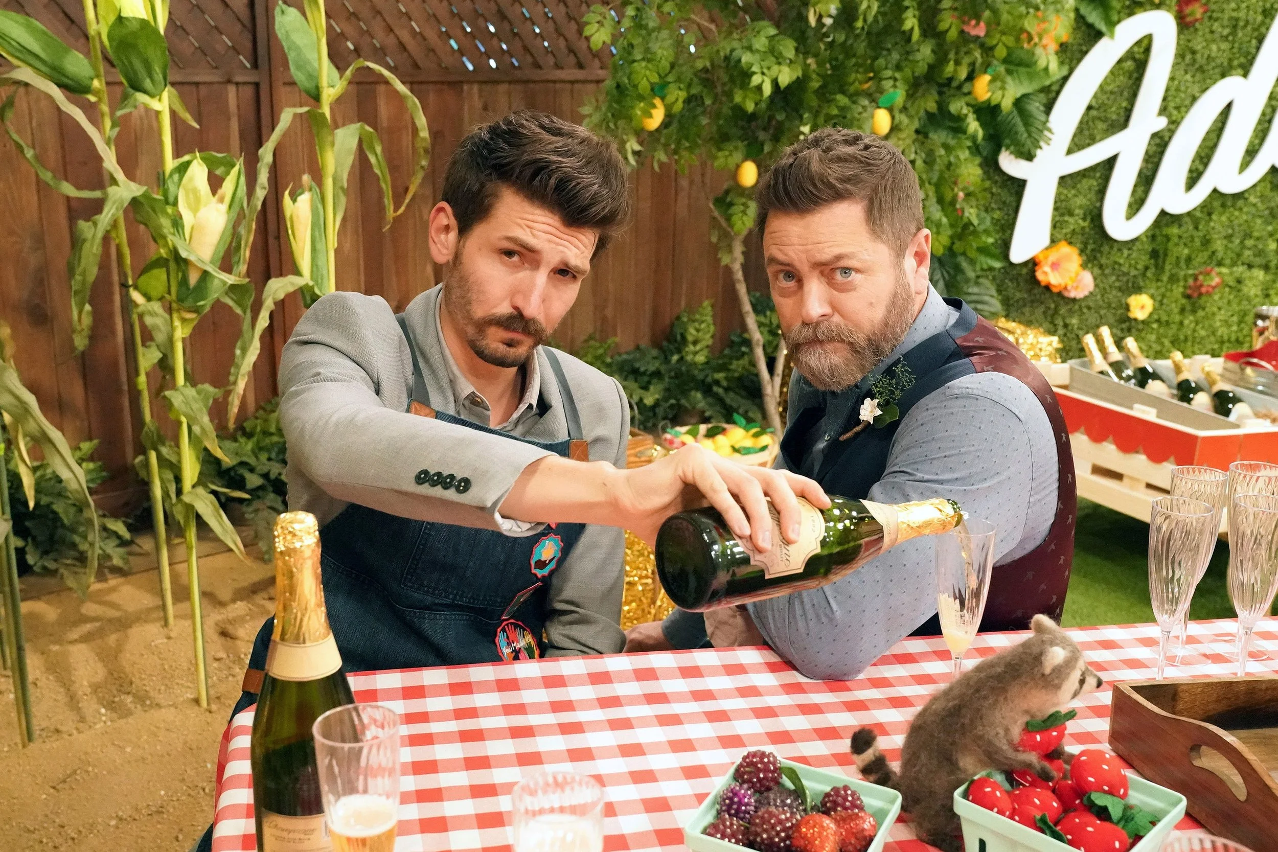 Two men are at a table with a red and white checkered tablecloth, pouring a bottle of champagne into glasses. The table has various berries and strawberries, and the background features greenery, lemon trees, and a wooden fence.