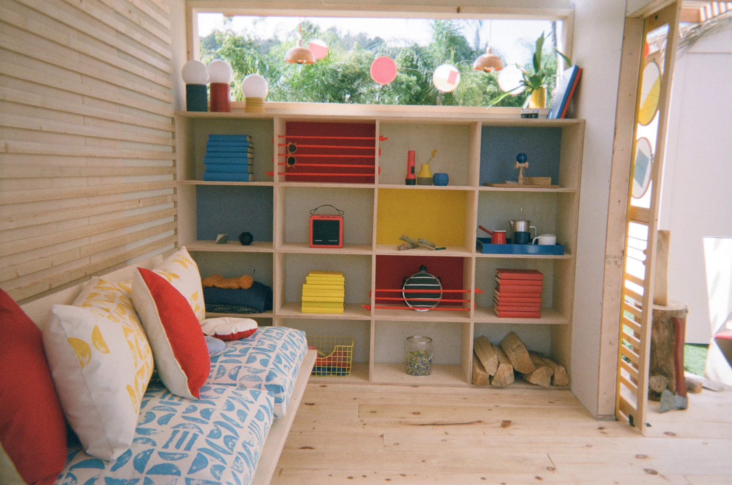 A colorful playroom with a large wooden bookshelf made of light-colored wood, decorated with toys, books, and storage baskets. There is a window behind the bookshelf showing green trees and blue sky outside.