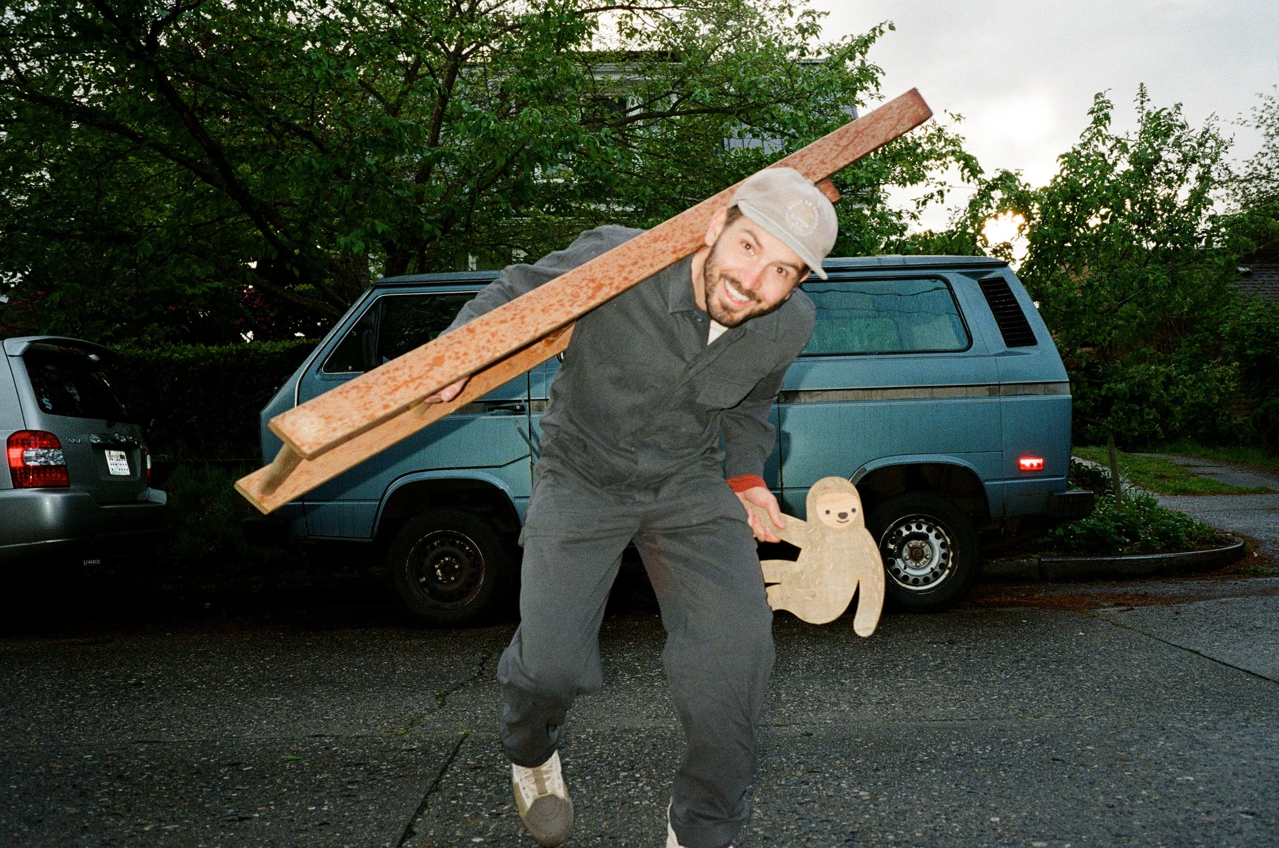 A man smiling and wearing a cap, carrying a large wooden plank on his shoulder, standing outdoors near parked cars and trees.