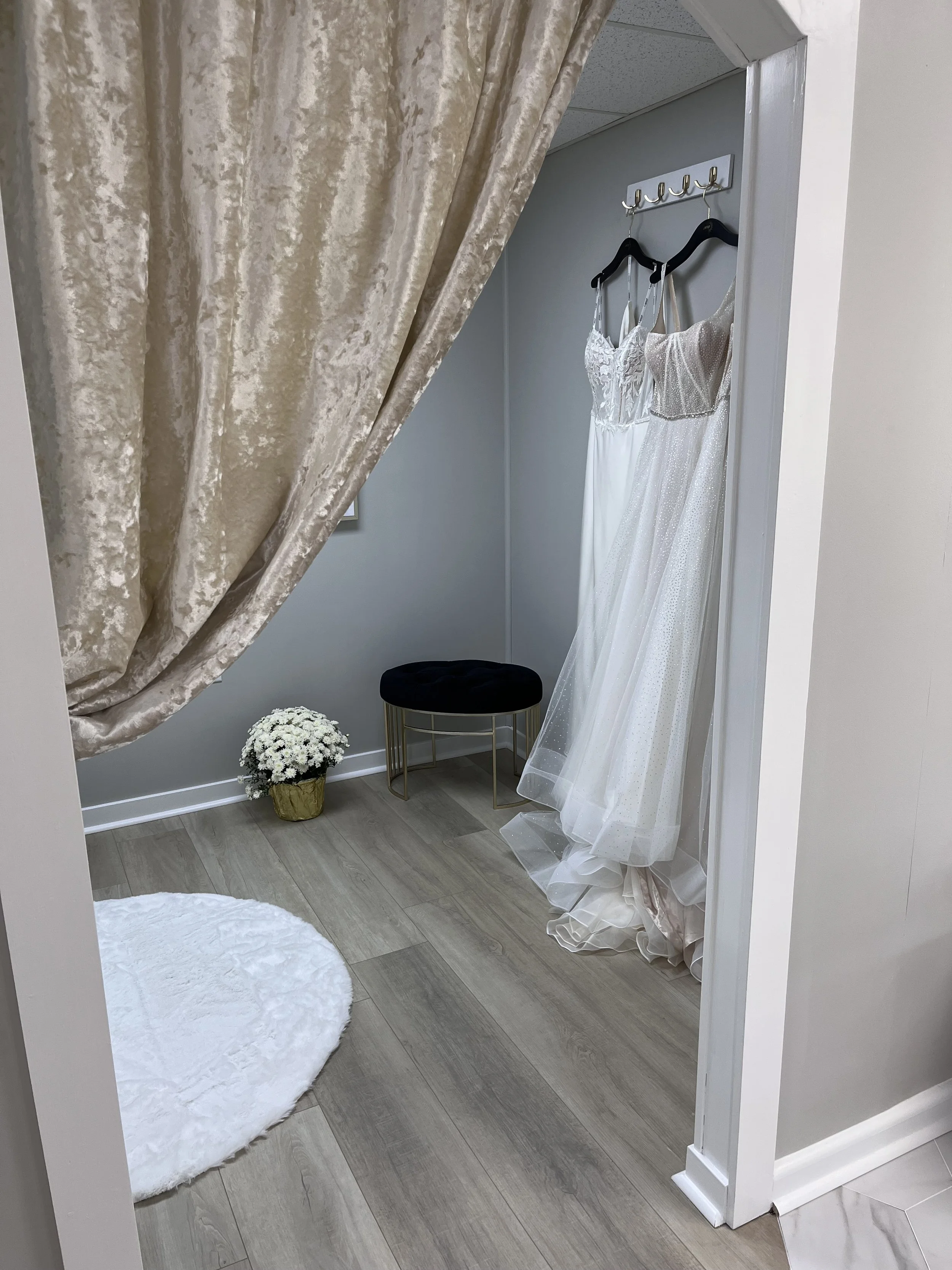 Two wedding dresses hanging on black hangers on a wall hook behind a beige curtain in a dressing room. A small round white rug on the floor, a potted white flower, and a small gold and black stool.