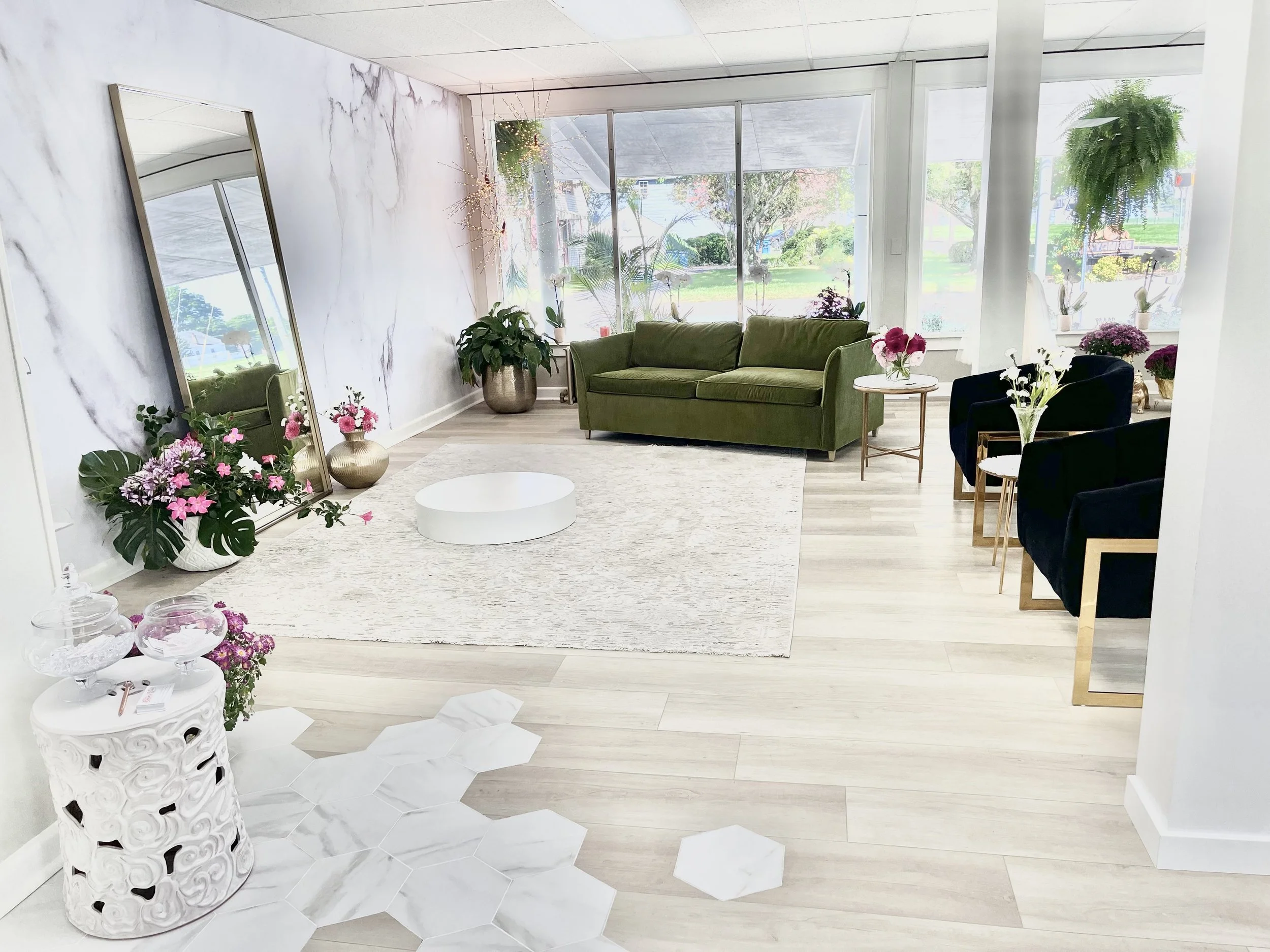 An interior living space with large windows, a green velvet sofa, black armchairs, and various pink and white flowers on tables and in pots. There is a tall gilded mirror leaning against the wall, a white intricately carved side table with floral arrangements, and paper decorations on the light-colored wood floor.