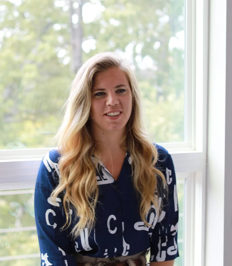 A woman with long blonde hair standing near a window with green trees outside, wearing a blue shirt with white lettering.