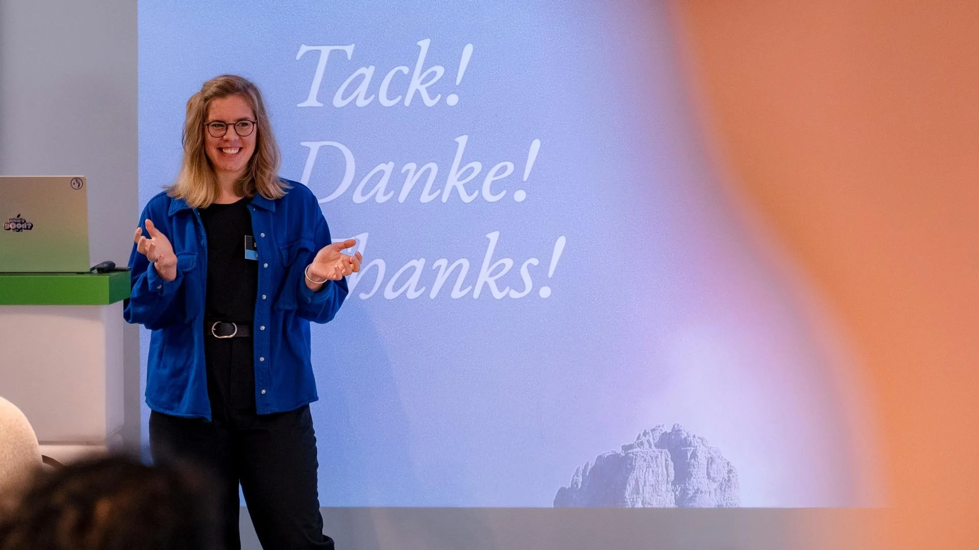 A woman with glasses and blonde hair smiling and gesturing with her hands in front of a presentation slide that says 'Tack! Danke! Thanks!' with a mountain illustration in the corner.