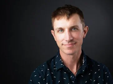 Portrait of a young man with short brown hair, blue eyes, wearing a dark blue shirt with small white patterns, against a dark gray background.
