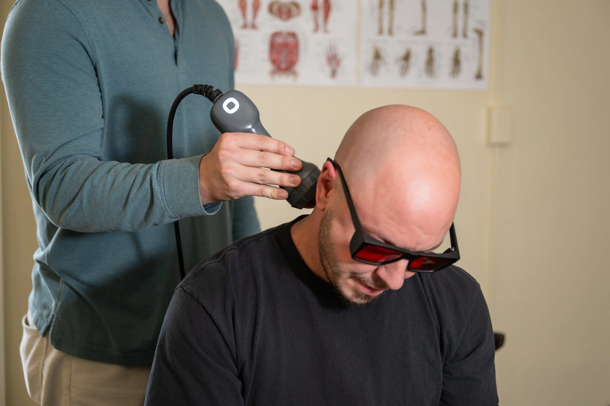 A person undergoing a medical or diagnostic procedure with a handheld device on his head, wearing protective glasses, while another person operates the device.