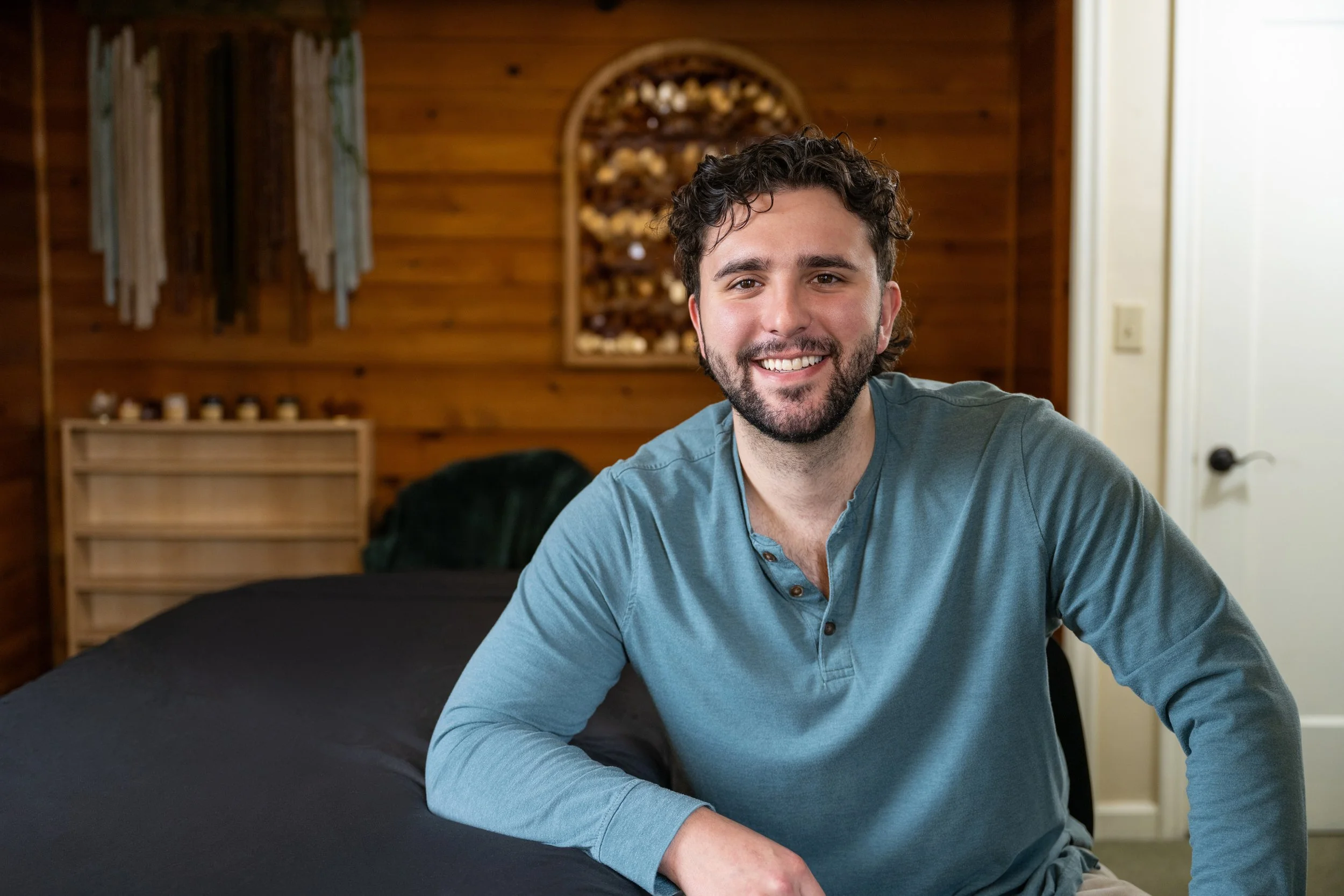 A smiling man with dark curly hair and a beard sitting on a bed in a wooden-paneled room.