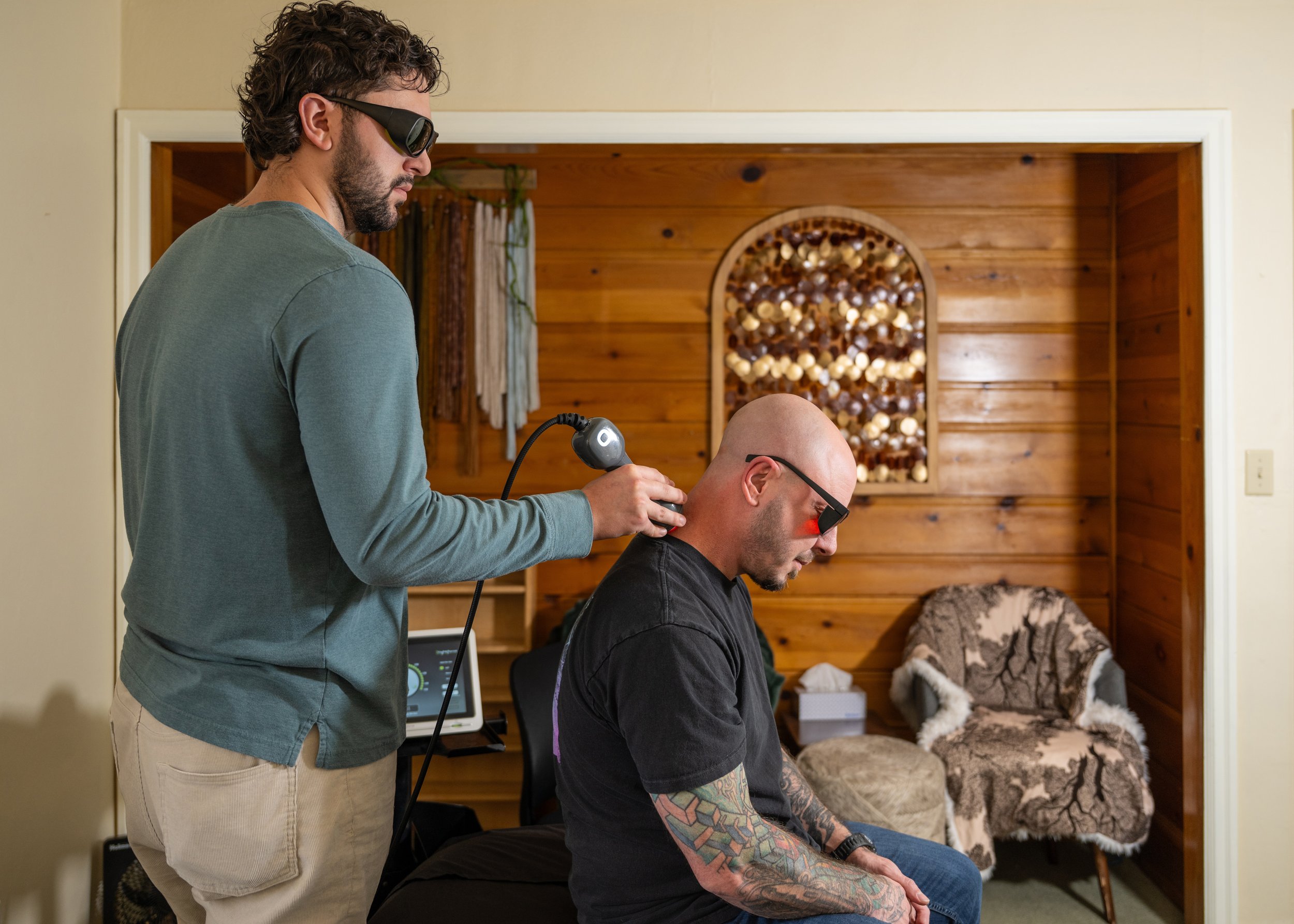 A man receiving a sonic massage therapy from a therapist in a cozy wooden-paneled room. The man is seated, wearing sunglasses, with tattoos on his arms, while the therapist stands behind him, wearing glasses and sunglasses, holding a sonic therapy device against his neck.