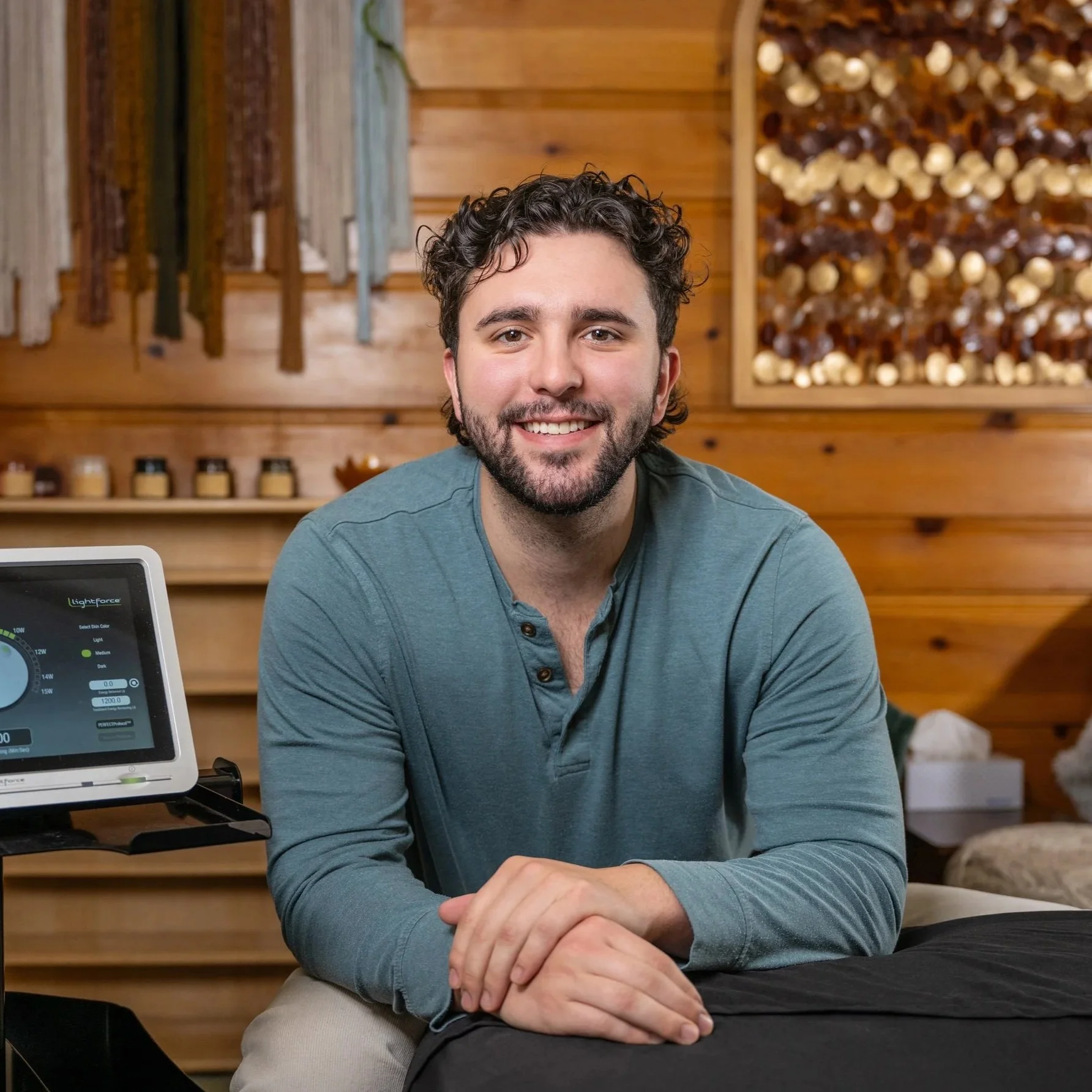 A smiling man with dark curly hair and a beard, wearing a teal long-sleeve shirt, sitting at a table in a wood-paneled room with decorative wood art on the wall.