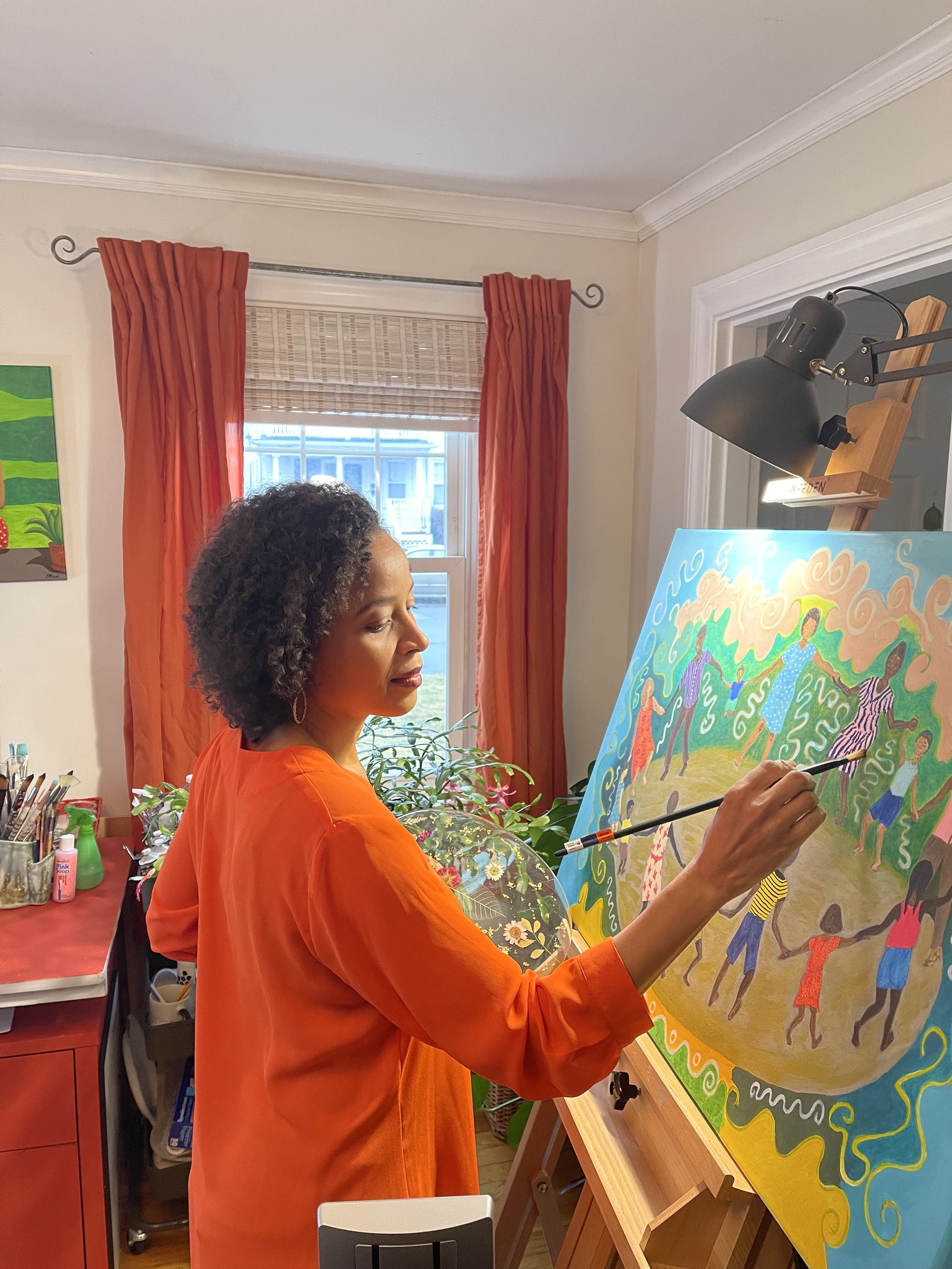 A woman with curly hair, wearing an orange top, is painting a colorful, intricate artwork on a canvas in a well-lit room. The room has cream-colored walls, orange curtains, and a window with blinds. There are art supplies and greenery around her.