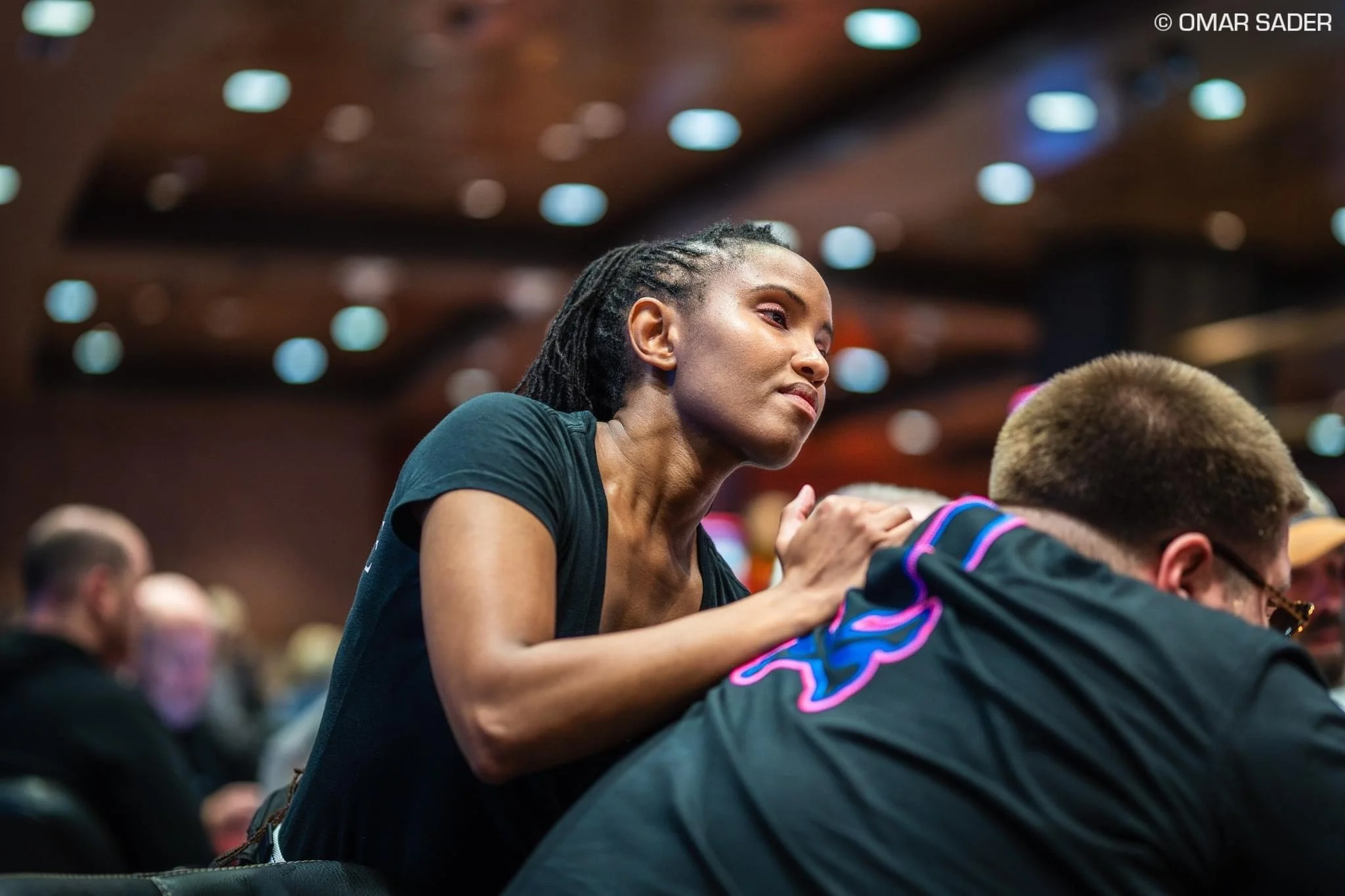 A woman with braided hair in a black shirt massaging a poker player with a furry hood in a dark indoor setting with blurred audience in the background.
