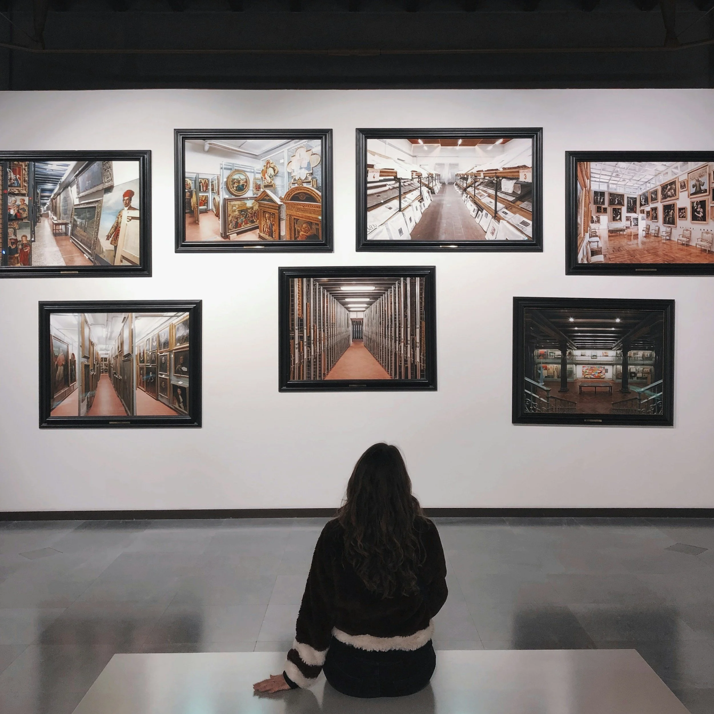Person with long hair sitting on a bench in an art museum, viewing framed artworks on a white wall.