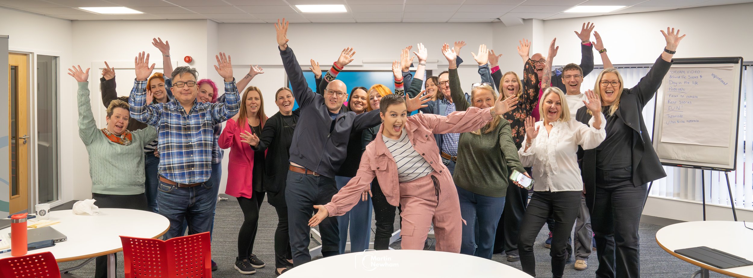 A group of people standing in an office or meeting room, smiling and raising their hands in celebration or excitement.