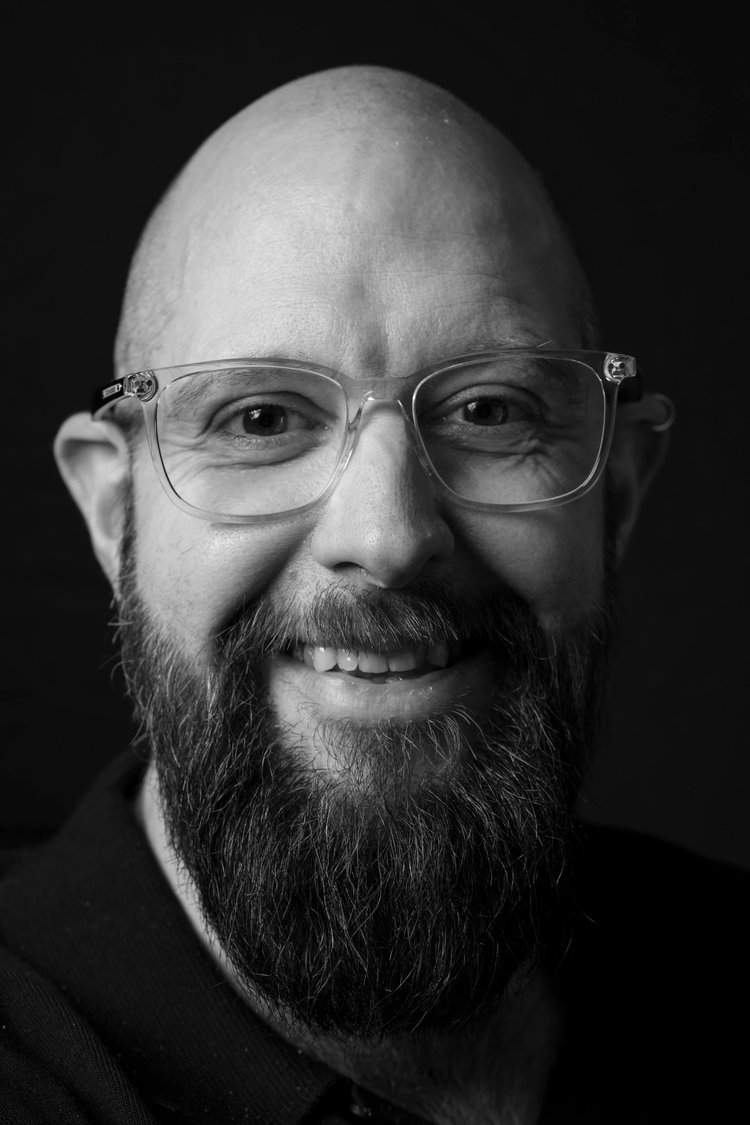 Close-up black-and-white photo of a smiling man with glasses, a beard, and a bald head.
