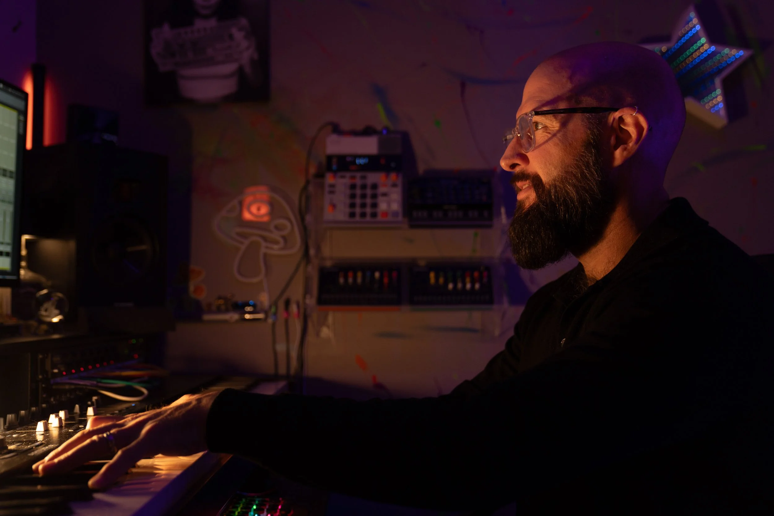 A man with glasses and a beard is playing a keyboard in a dimly lit room filled with music equipment and colorful LED lights.