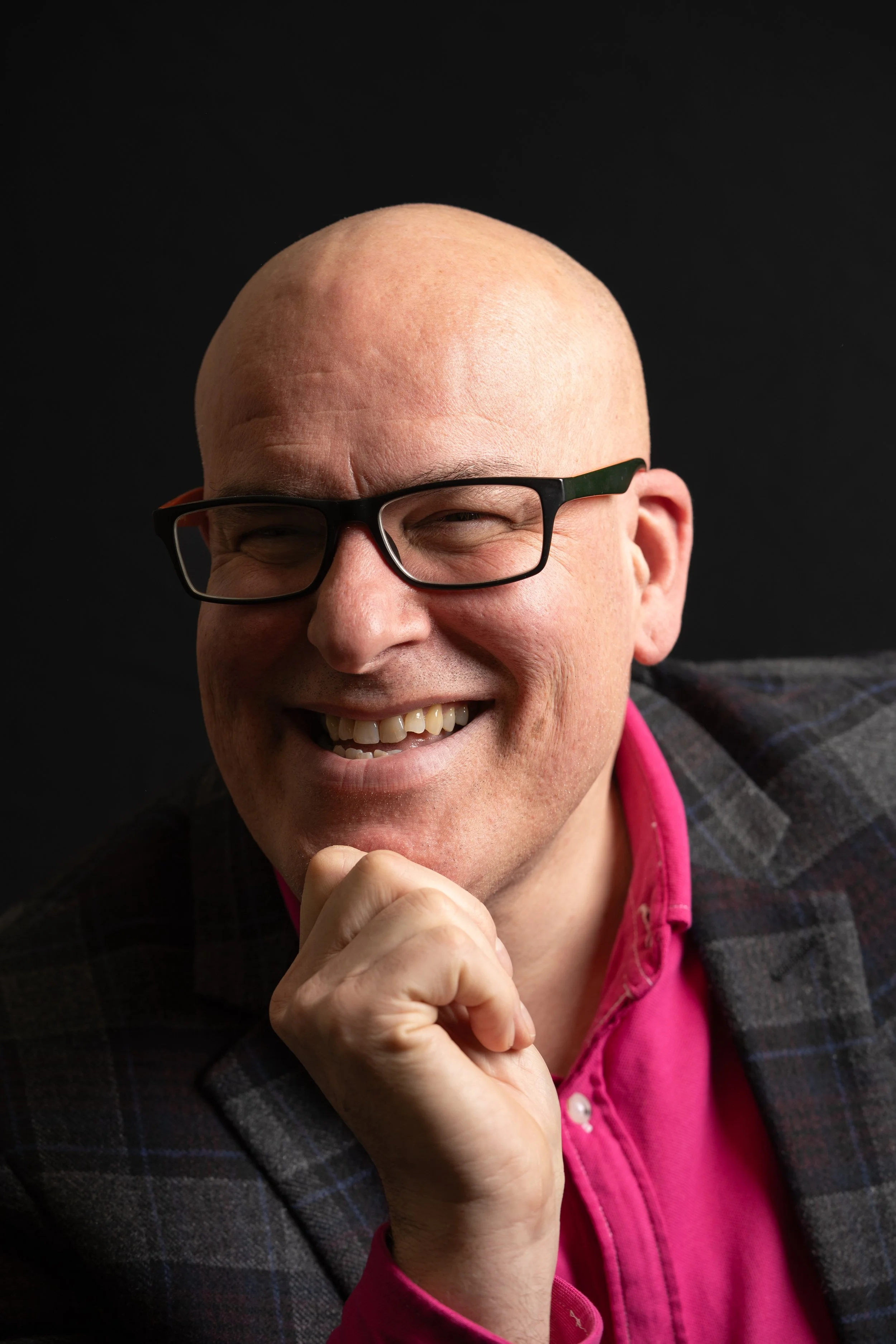 Portrait of a smiling, bald man wearing glasses, a pink shirt, and a plaid blazer, with his hand resting on his chin.