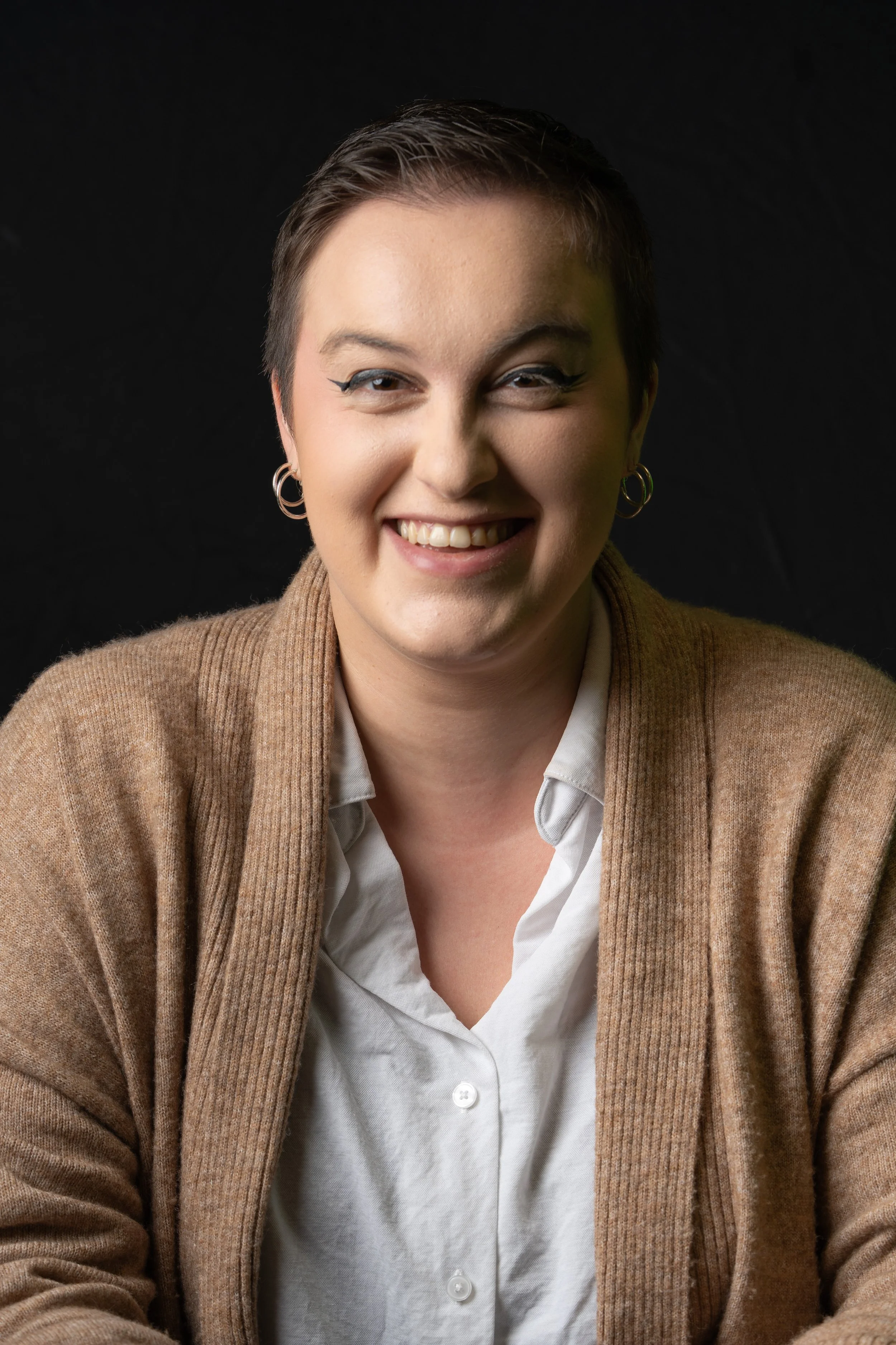 A woman with short hair, hoop earrings, and eyeliner smiling in front of a black background, wearing a white shirt and beige cardigan.