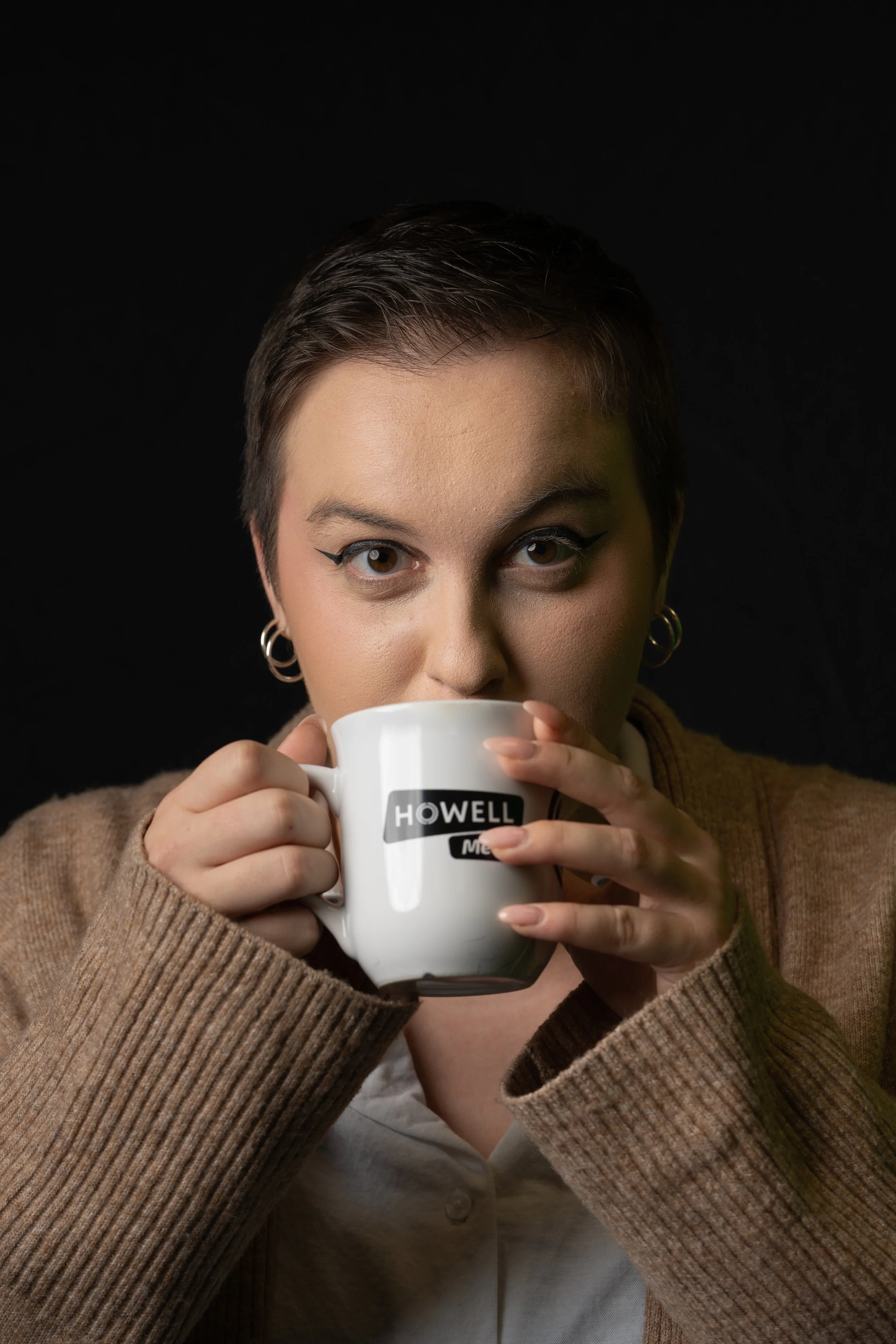 A woman with short brown hair and hoop earrings holding a white mug with black text that reads 'HOWELL' in front of her face against a dark background.