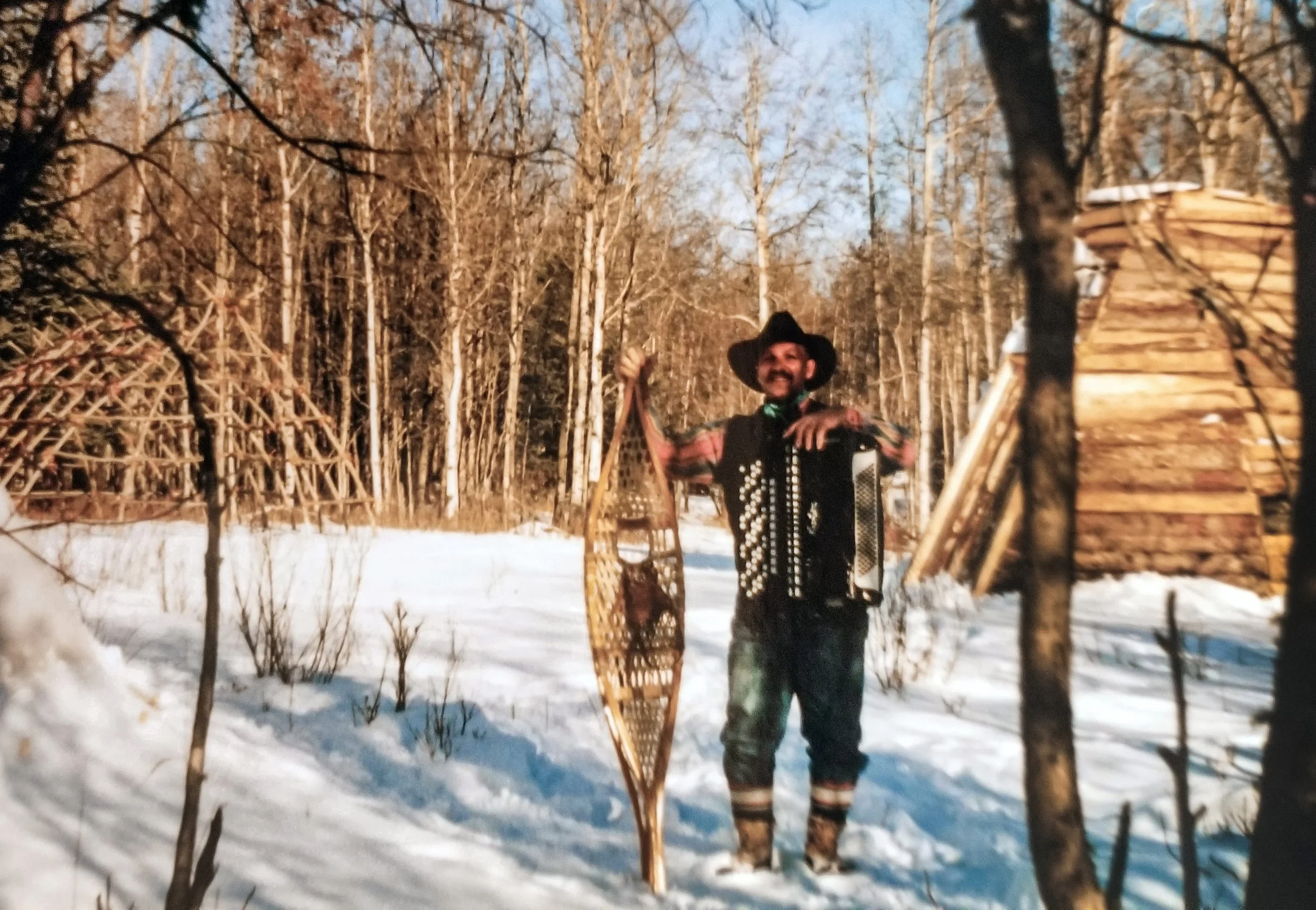 A man wearing traditional clothing and a cowboy hat smiling while holding a snowshoe and an accordion, standing in a snowy forest with bare trees and a wooden structure in the background.