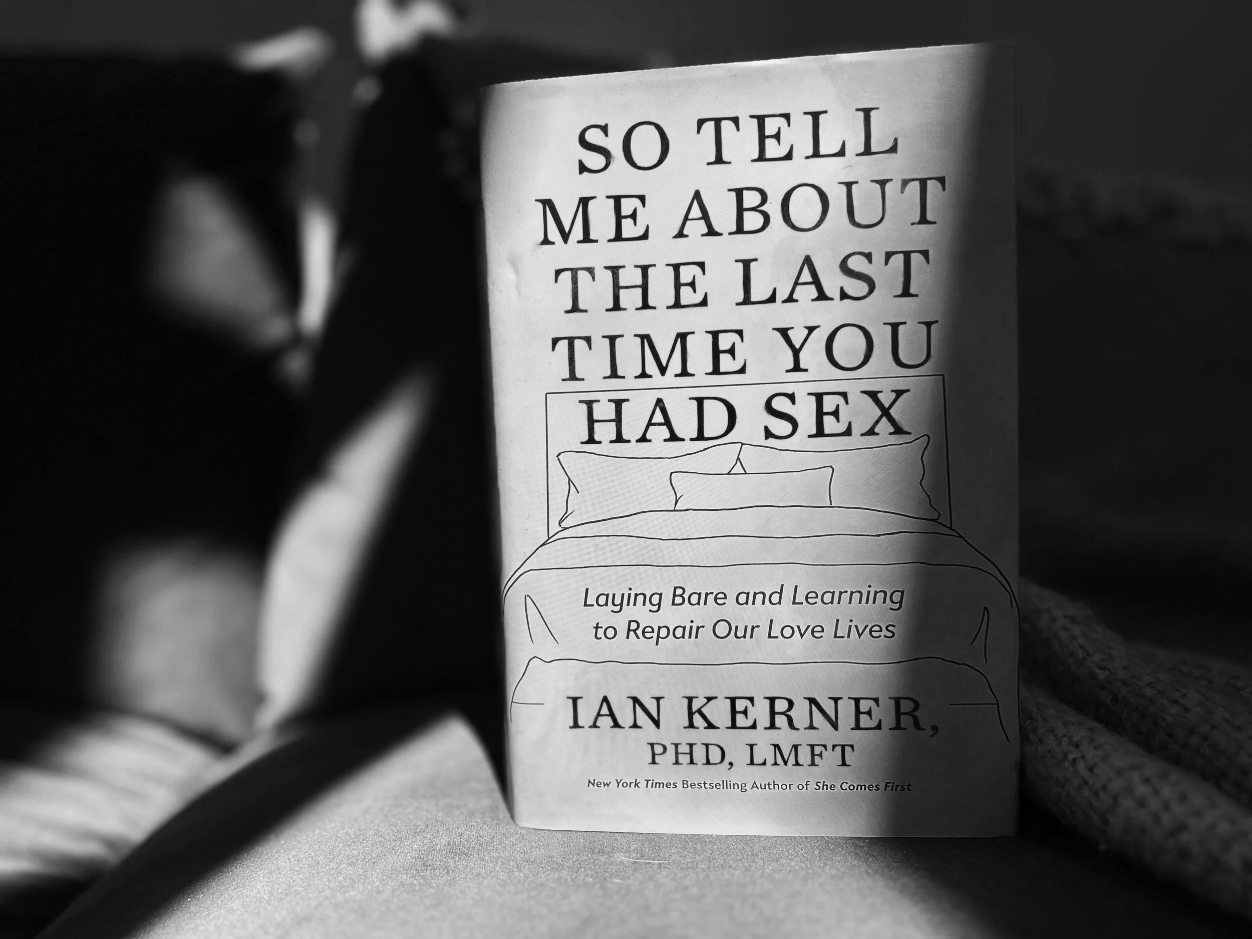 Black and white photo of a book with the title, "So Tell Me About the Last Time You Had Sex" by Ianky Kerner, PhD, LMFT, and a subtitle about laying bare and learning to repair love lives, with blurred objects surrounding it.