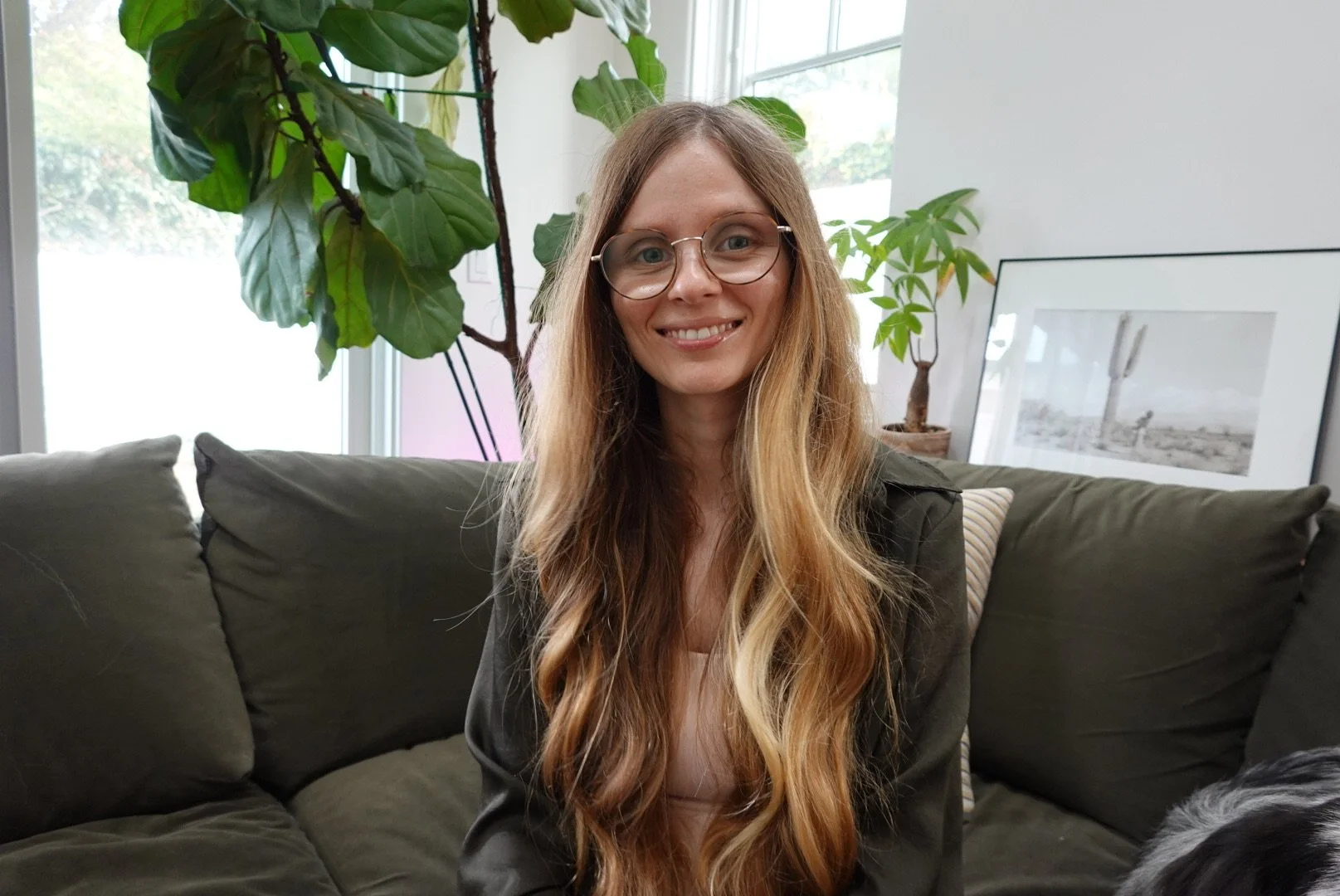 A woman with long, wavy blonde hair and glasses smiling and sitting on a dark green couch in a living room with green plants and framed artwork in the background.