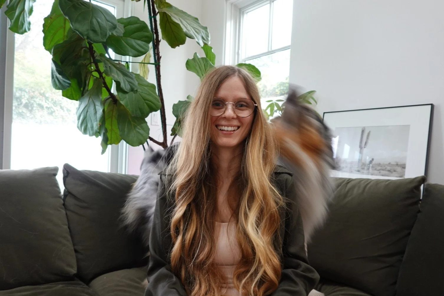 A woman with long wavy blonde hair and glasses smiling while sitting on a dark green sofa in a bright living room with large plant leaves and a framed photograph on the wall behind her, with a mink-like animal blurred in motion next to her.