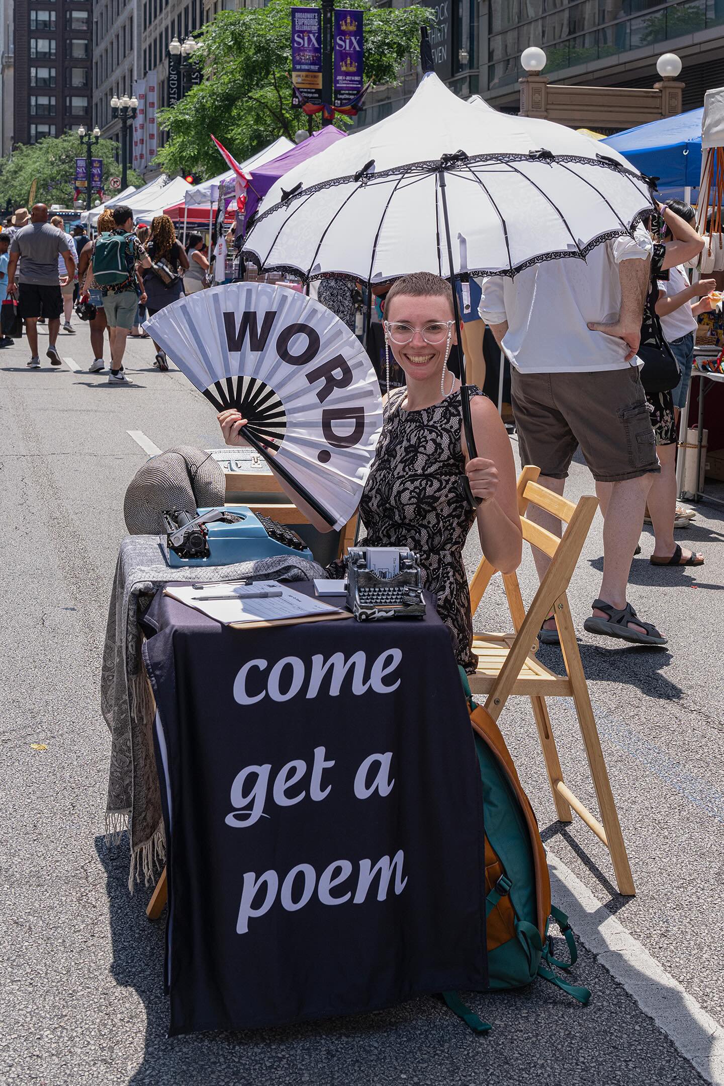 Street scene during a public event with multiple tents and people walking. A woman in the foreground is smiling and holding a fan with the words 'WORD,' 'to,' 'get,' 'a,' and 'poem' visible. She is seated at a table with a typewriter and papers. The woman is wearing glasses and a floral dress. An umbrella shades her. The background shows more tents, trees, and urban buildings, with banners hanging from street lamps, indicating a festival or market.