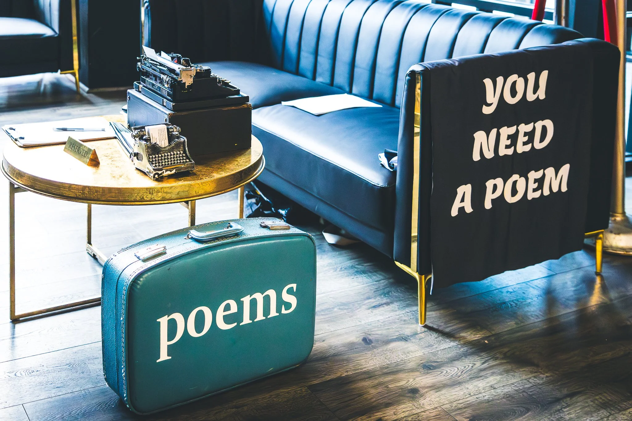 Vintage typewriter on a black box, a reserved sign on a round gold table, a blue suitcase, and a teal suitcase with 'poems' written on it, in front of a black sofa with white writing that says 'you need a poem' in a cozy room.