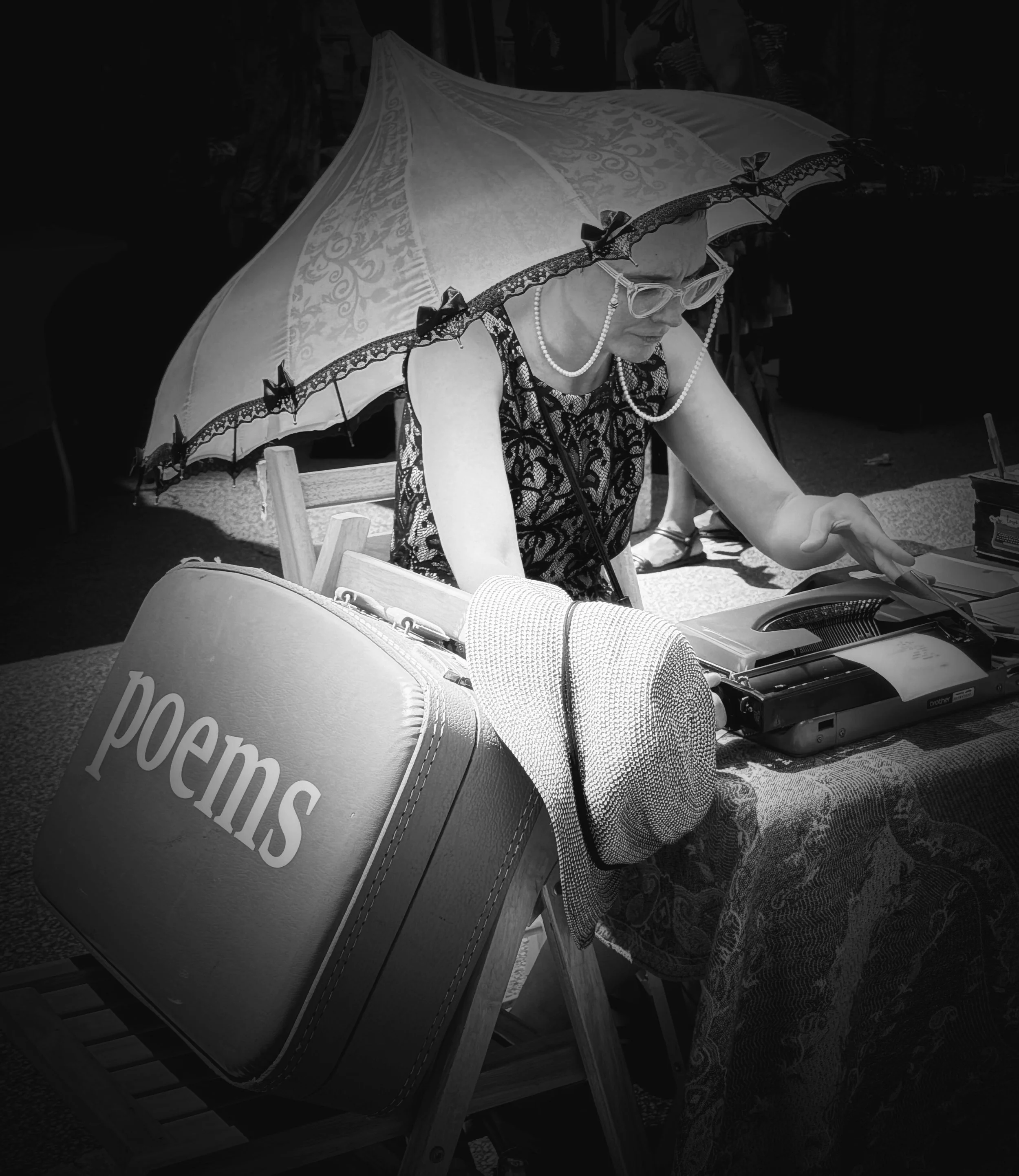 A woman wearing glasses, a floral dress, and pearl necklaces sitting under a large decorative parasol at an outdoor event, with a popcorn bag in the foreground.