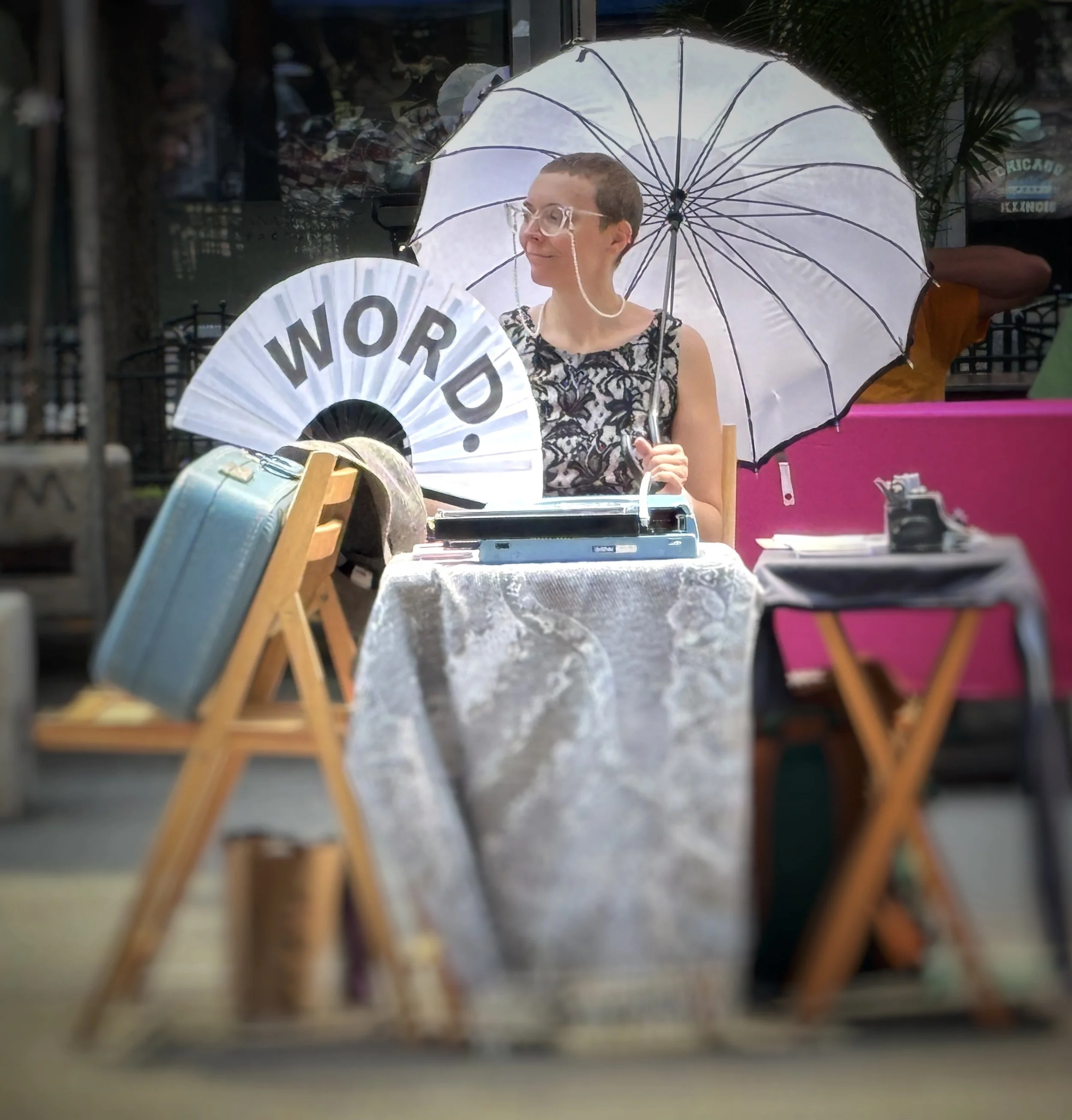 A woman smiling while holding a white umbrella with black ribs. She is sitting at a table with various items and a large paper fan with the word 'WORD?' written on it. The setting appears to be inside a shop or market with shelves and clothing in the background.