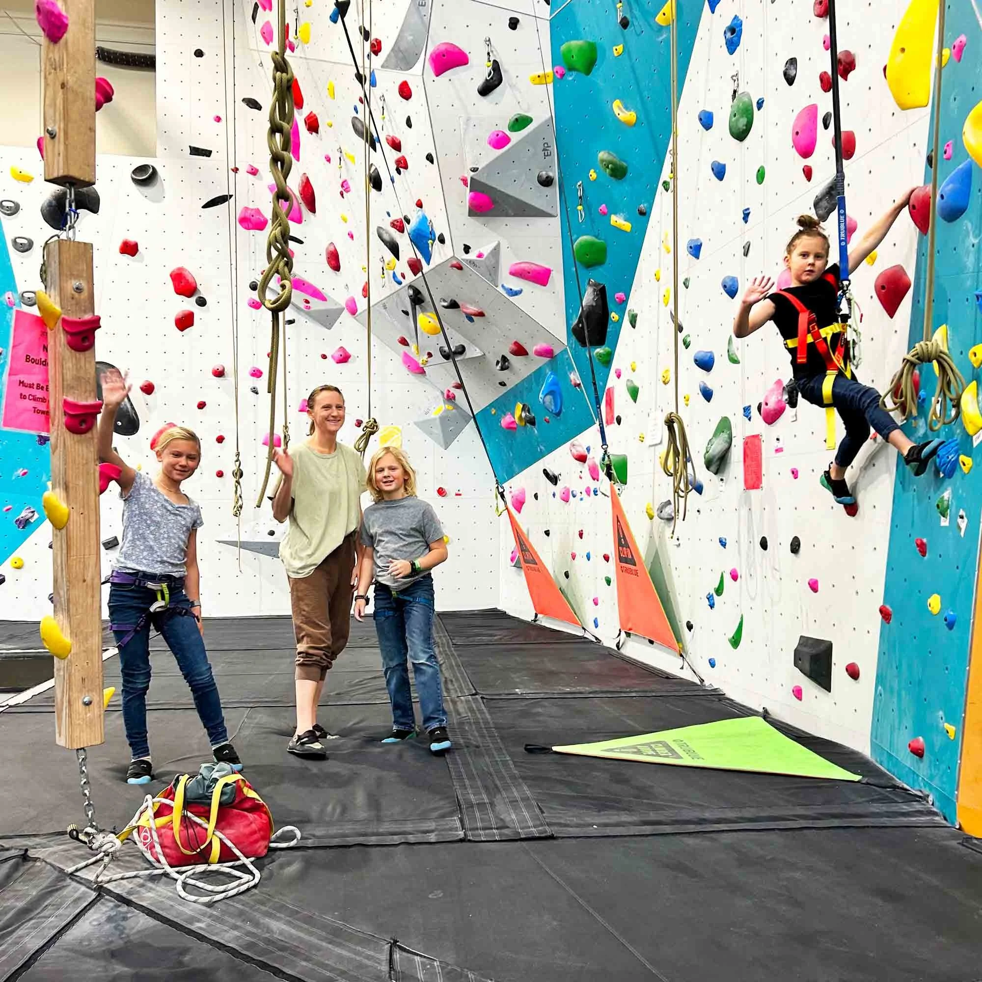 Three children and a woman at an indoor climbing gym. One girl is climbing the wall while the other two stand on the ground, smiling and waving. The wall has colorful holds and large geometric climbing structures.