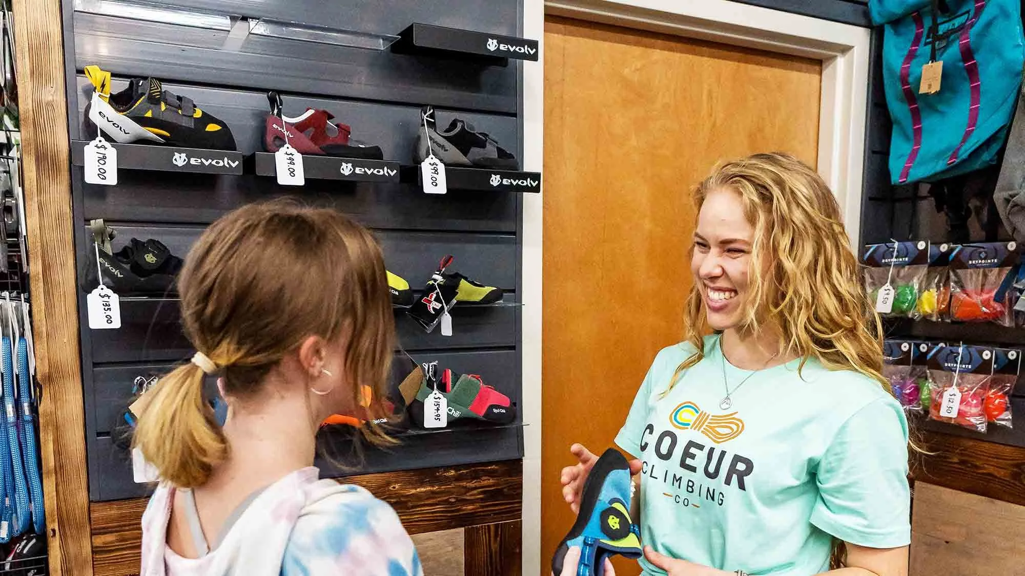 One woman consulting about climbing shoes with Coeur Climbing staff, and climbing shoes displayed on shelves behind them.