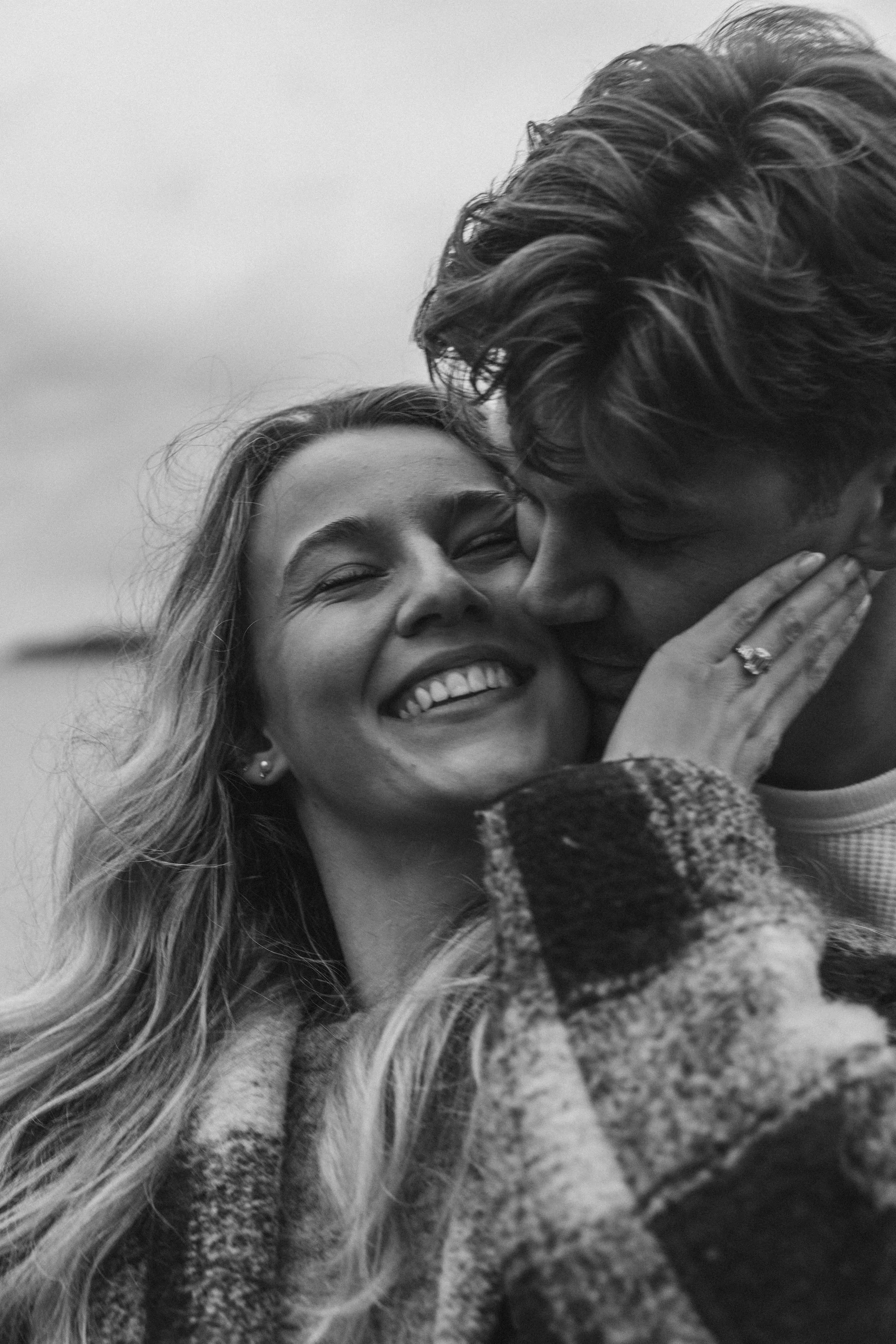 A joyful woman with long hair and a ring on her finger embracing and smiling as a man kisses her on the cheek.