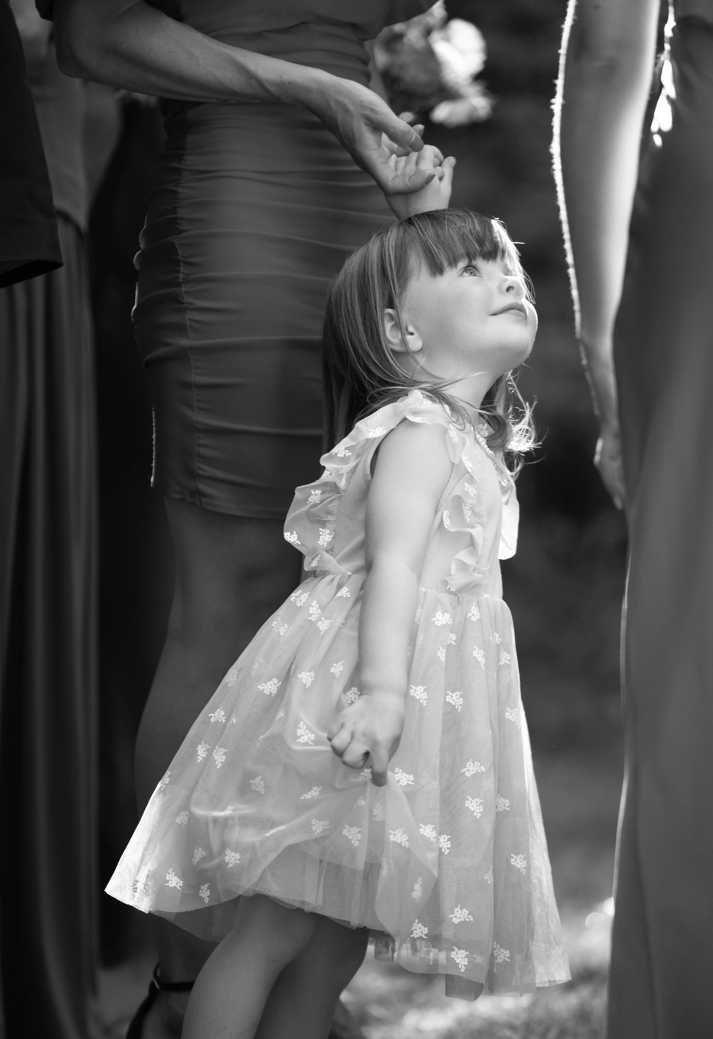 A young girl in a frilly dress holds hands with an adult, looking upward with a smile, outdoors during daytime.