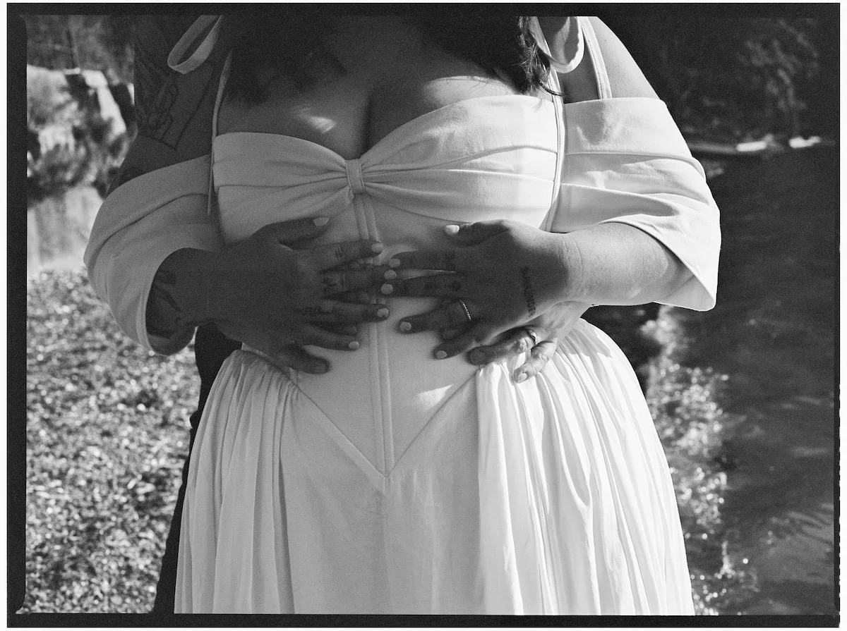A bride in a strapless wedding dress being embraced from behind by another person, with hands on her stomach and waist, near a body of water. Coast and Kin Wedding Photographer BC Wedding Photographer, Salt Spring Island Wedding Photographer